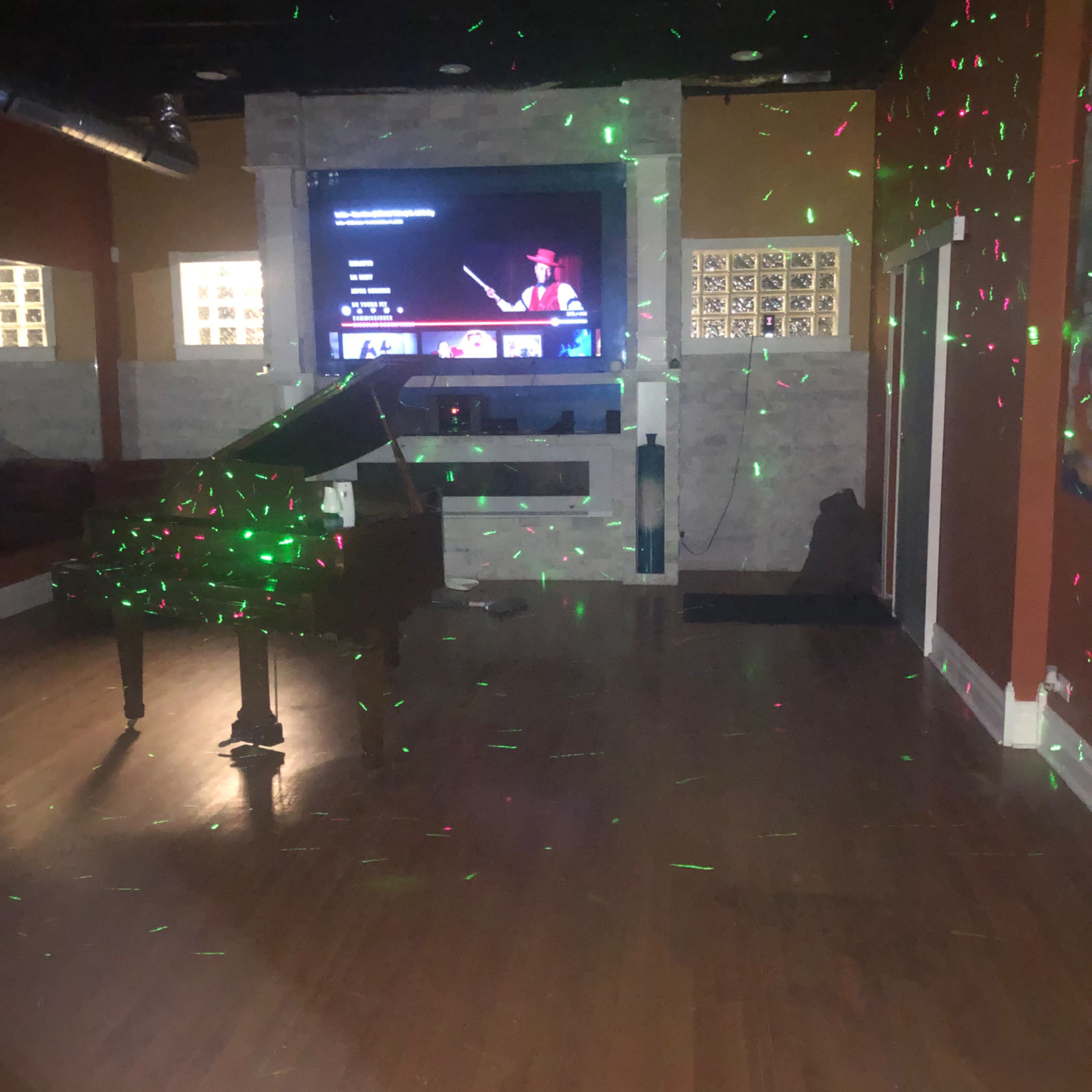 A room with wooden flooring, a grand piano in the center, and a large television displaying content on the wall, illuminated by green and red laser lights.