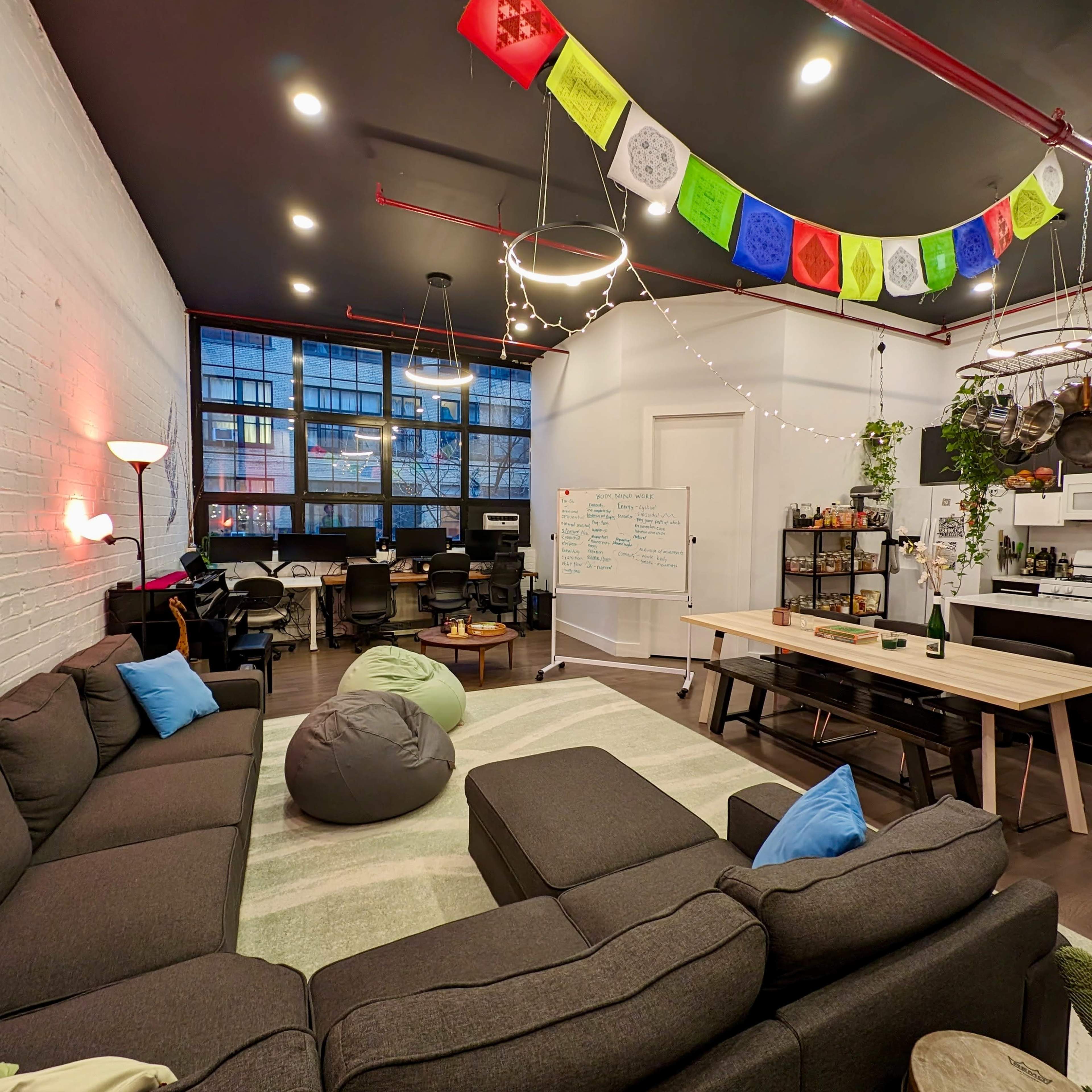 A modern, open office space with a sectional sofa, bean bags, a long dining table, and colorful banners hanging from the ceiling.