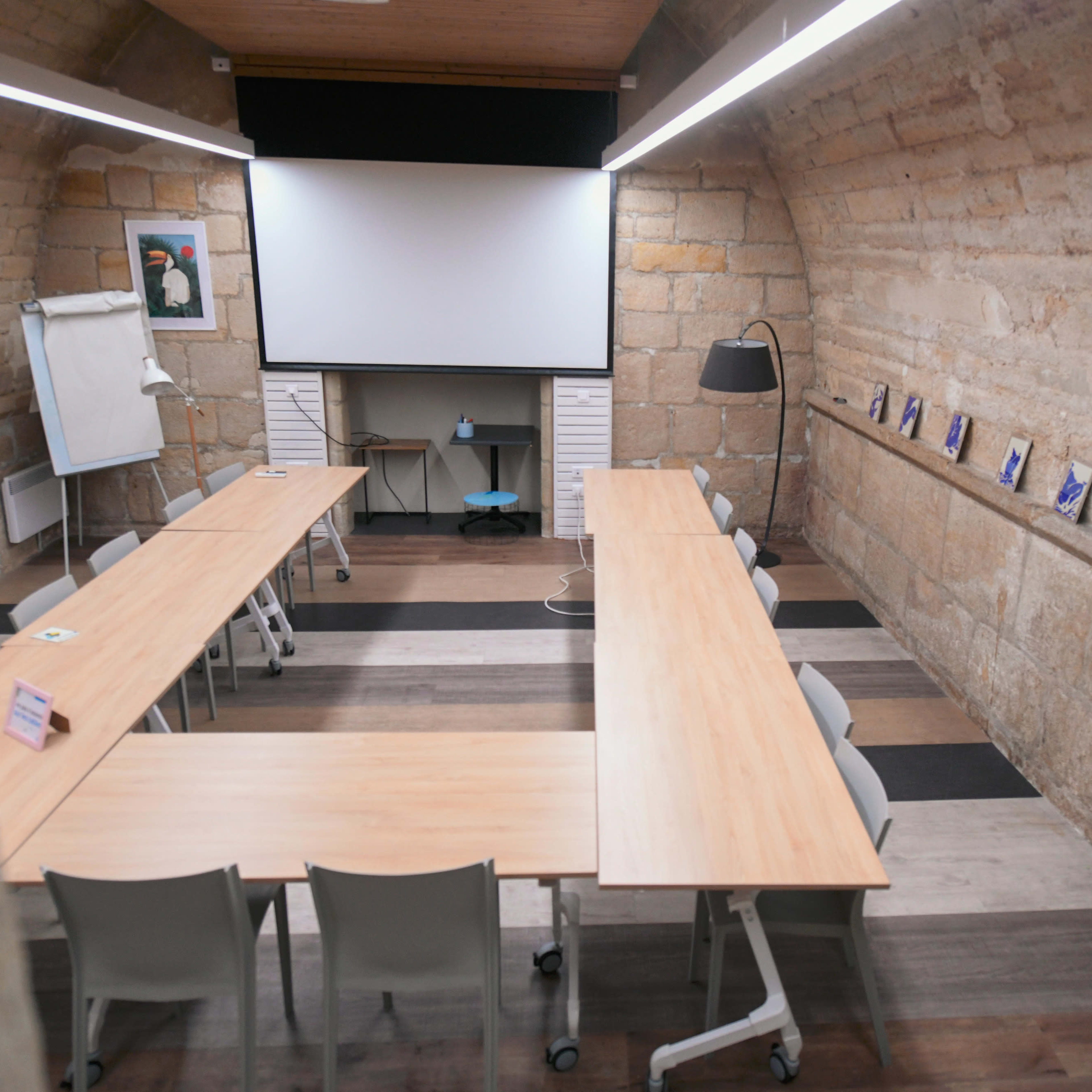 An empty meeting room with wooden tables arranged in a U shape, a large screen at the front, and stone walls.