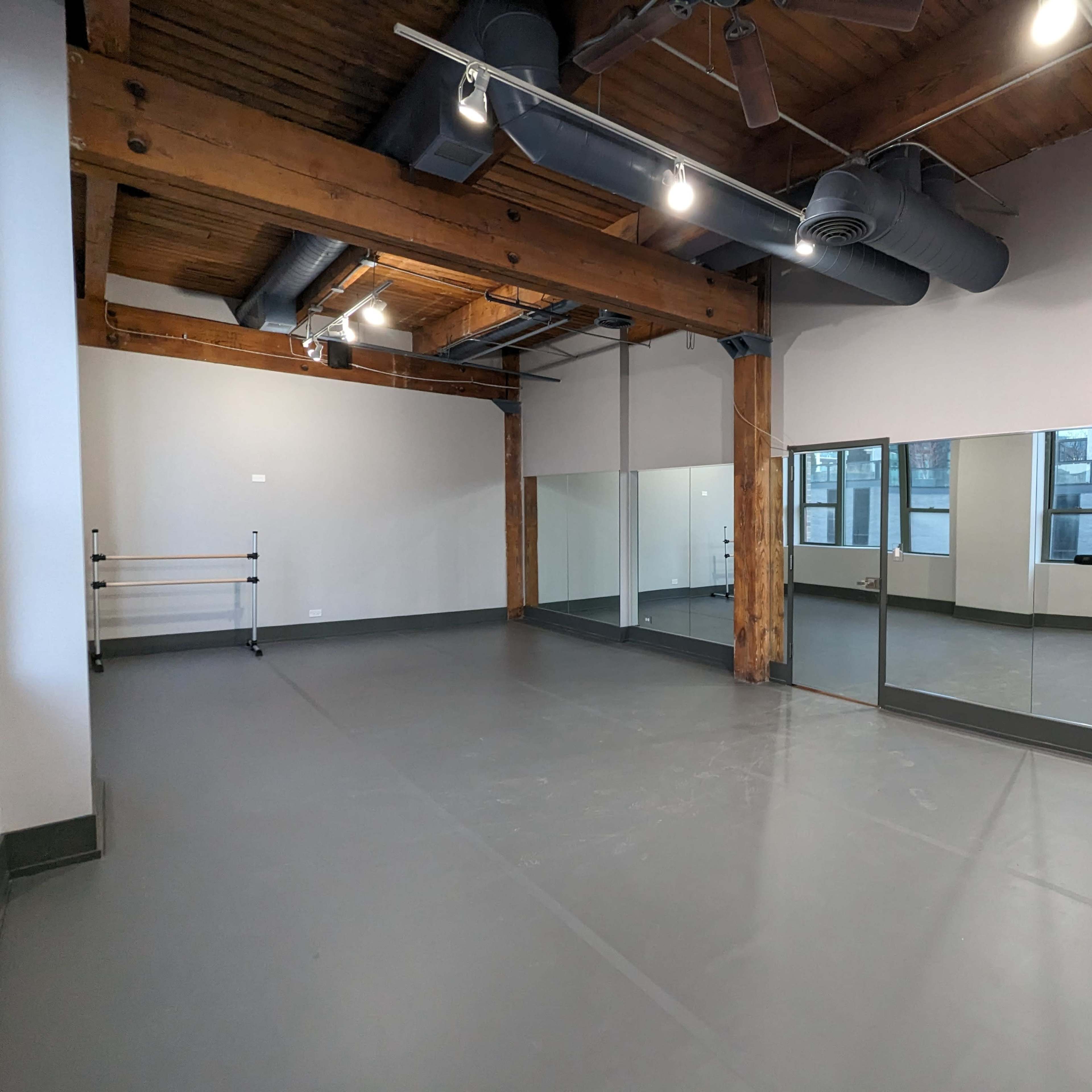 An empty studio space with wooden beams, large mirrors on one wall, and a gray floor.