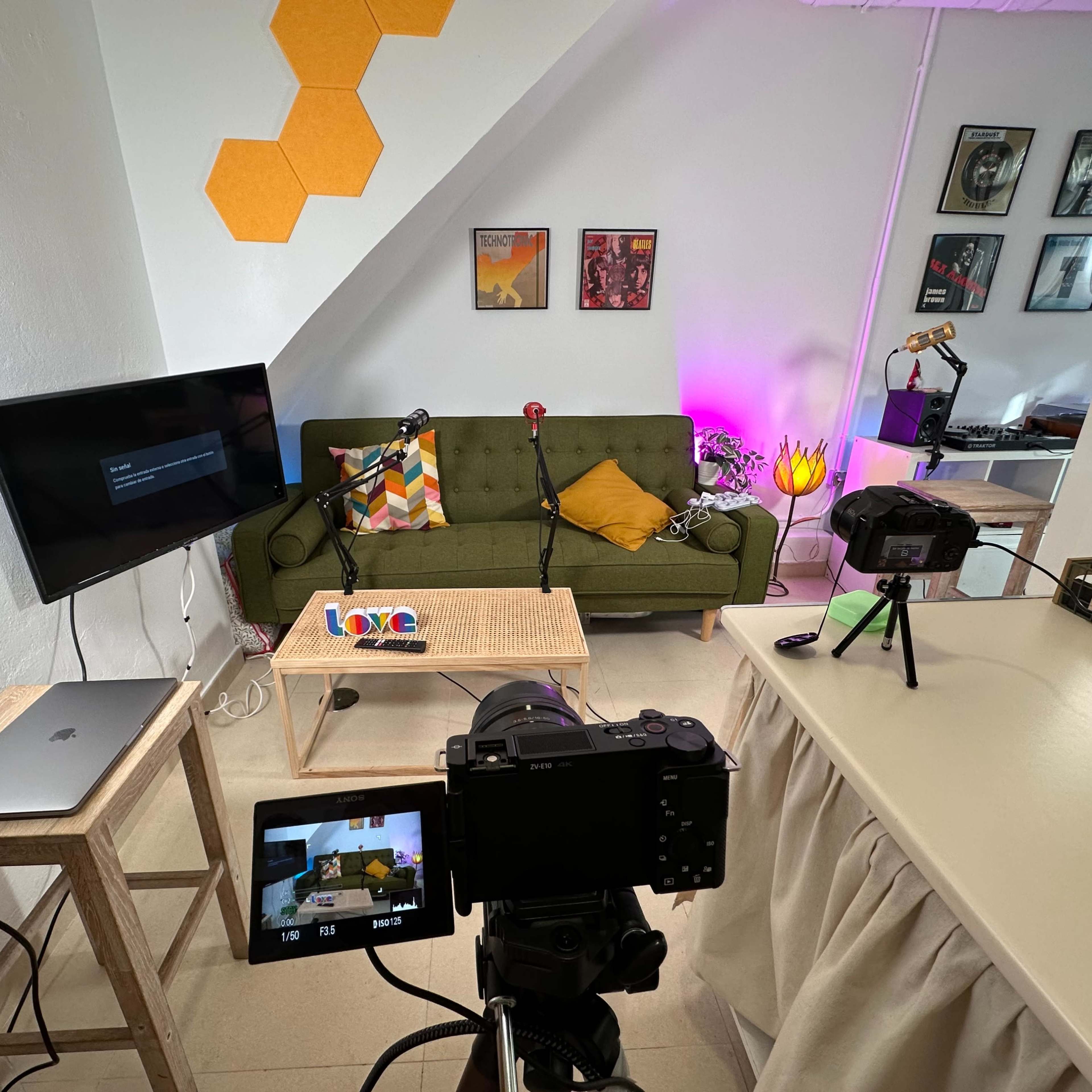 A camera and laptop are set up in a well-lit room featuring a green sofa, a television, and colorful wall decorations.