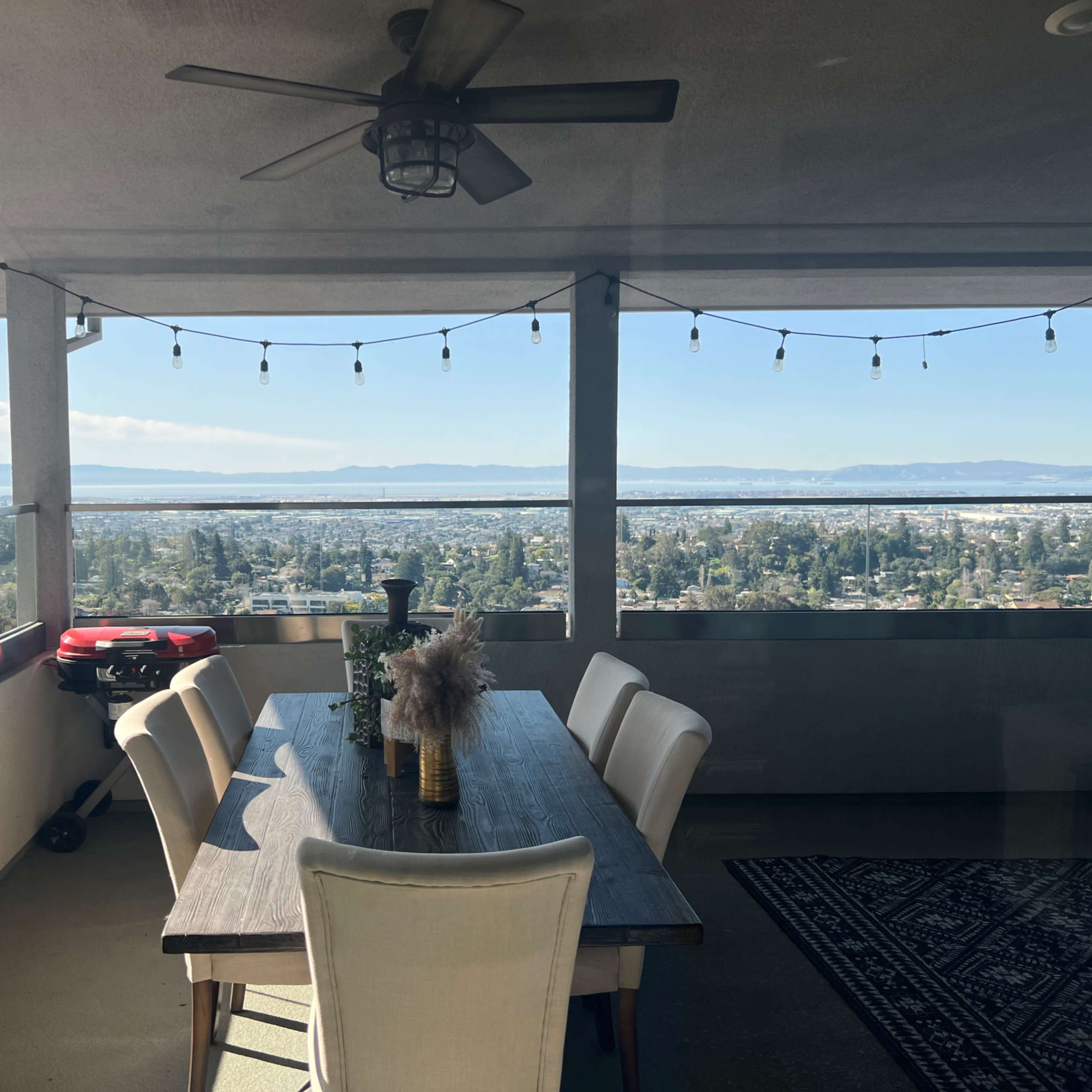 A dining table with six chairs is set on a balcony overlooking a cityscape and distant mountains, with string lights hanging above.