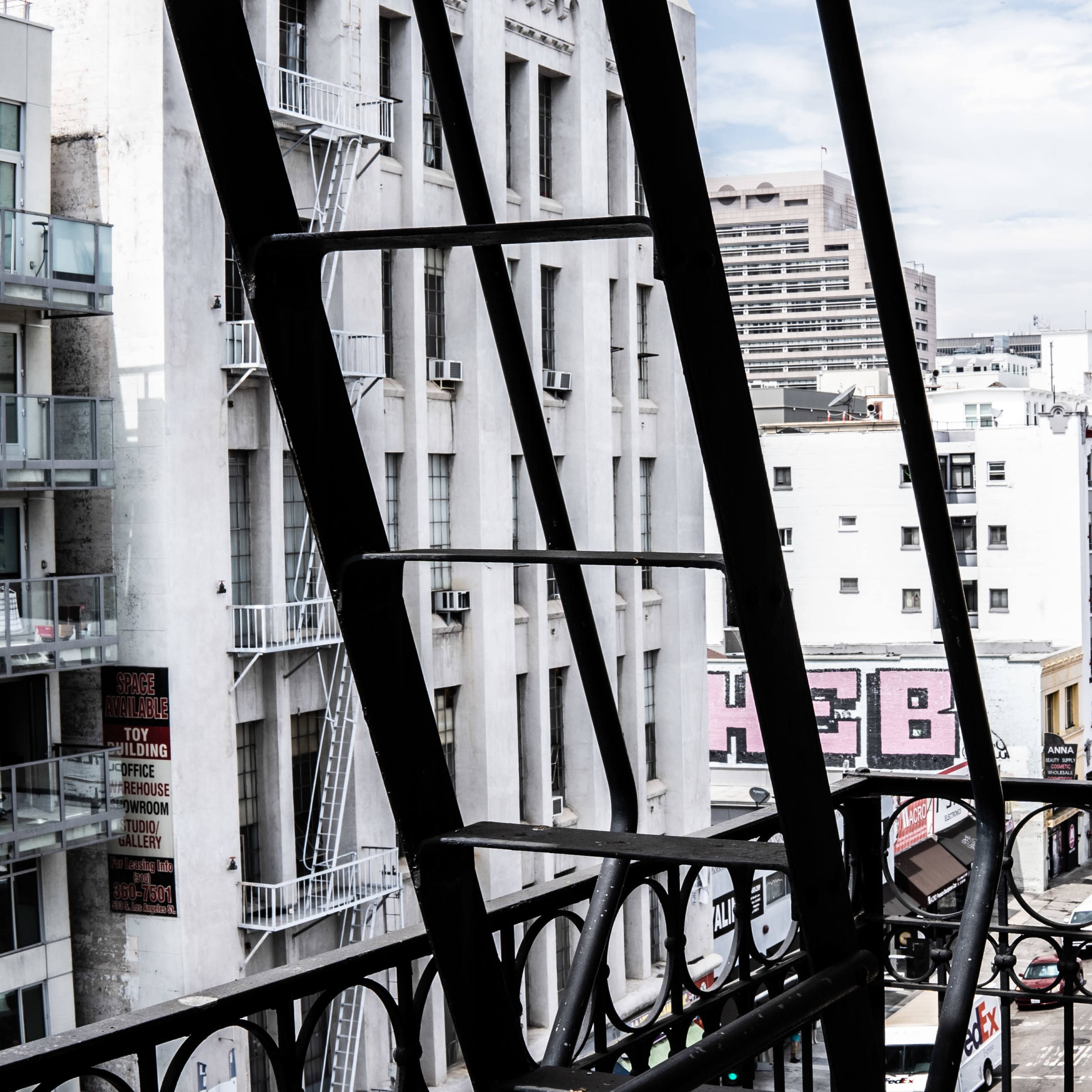 A view of a city street lined with buildings, framed by an iron fire escape on the left.