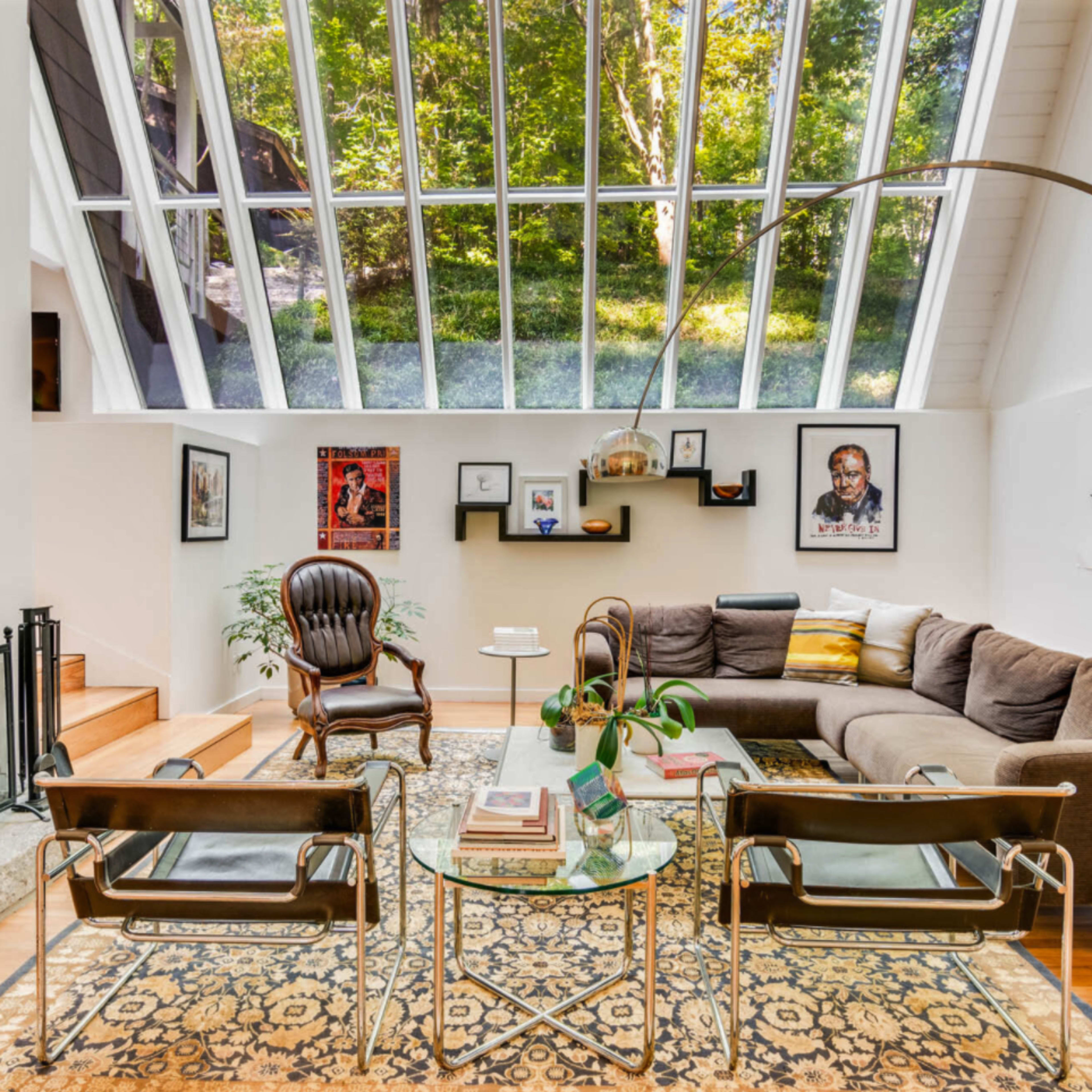 A modern living room with large glass windows, a sectional sofa, an armchair, a fireplace, and various framed artworks on the walls.