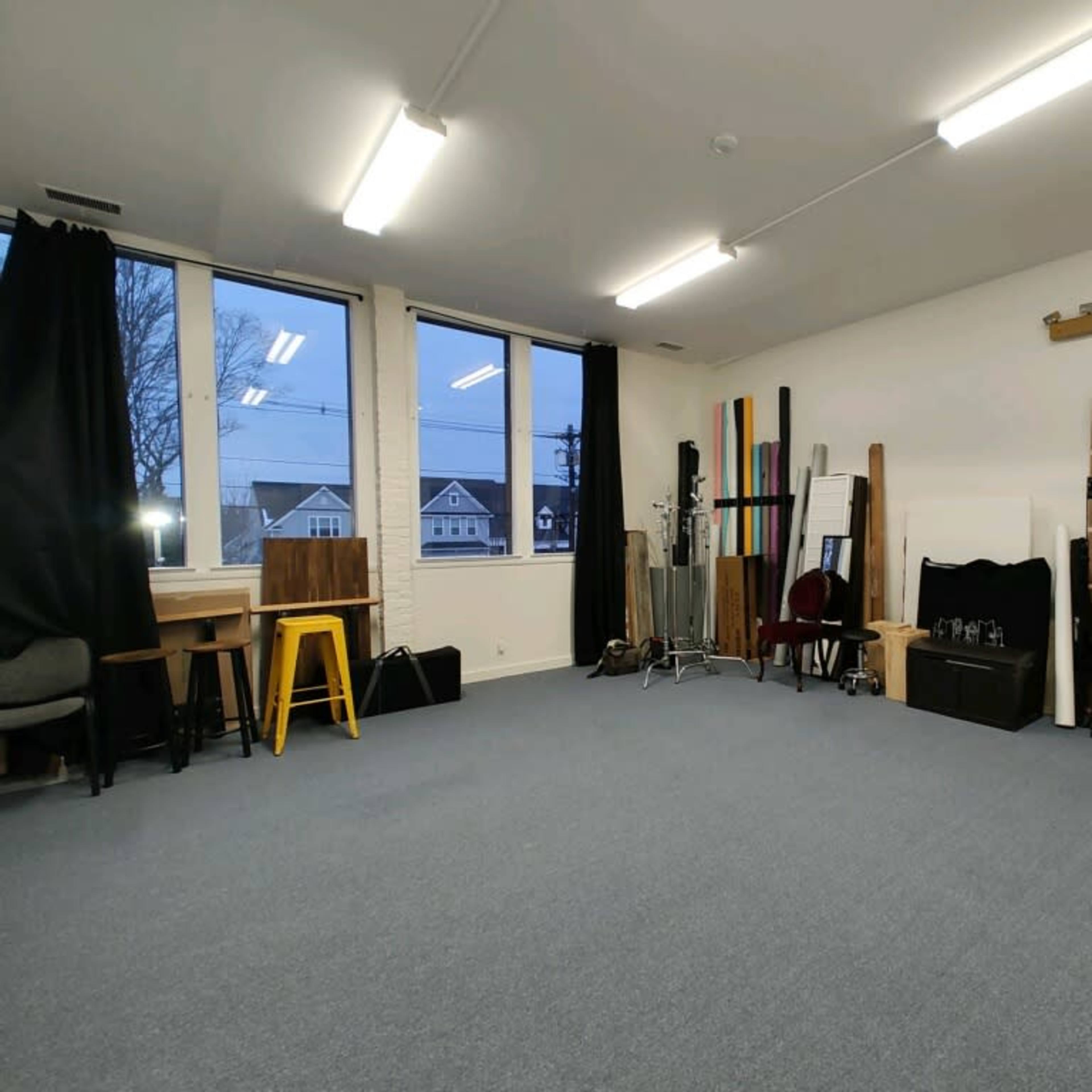 A spacious interior studio with a carpeted floor, large windows, and various rolls of colored backdrops neatly arranged against the wall.