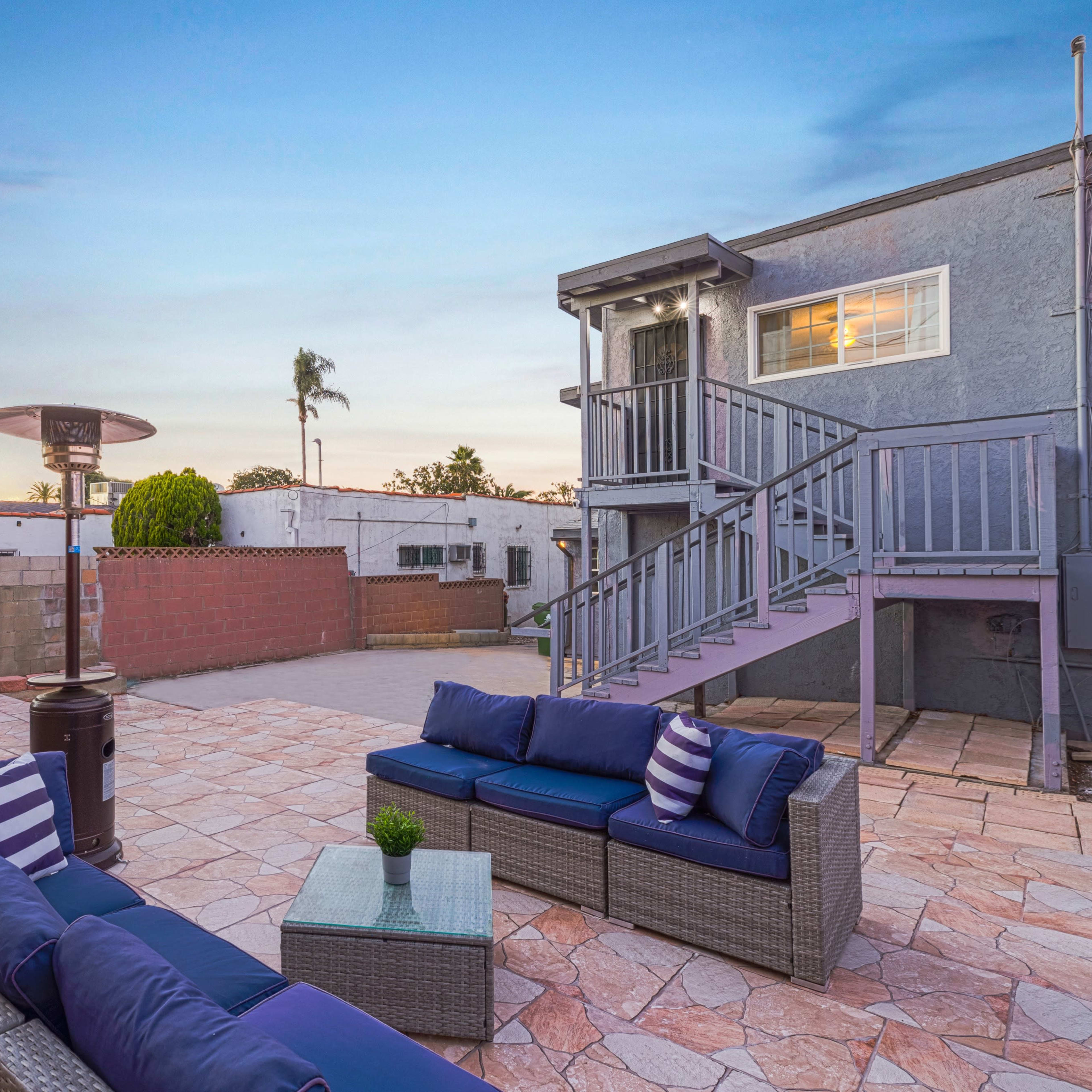 A backyard patio with a seating area, outdoor heater, and a multi-station gym equipment set against a background of a two-story building.