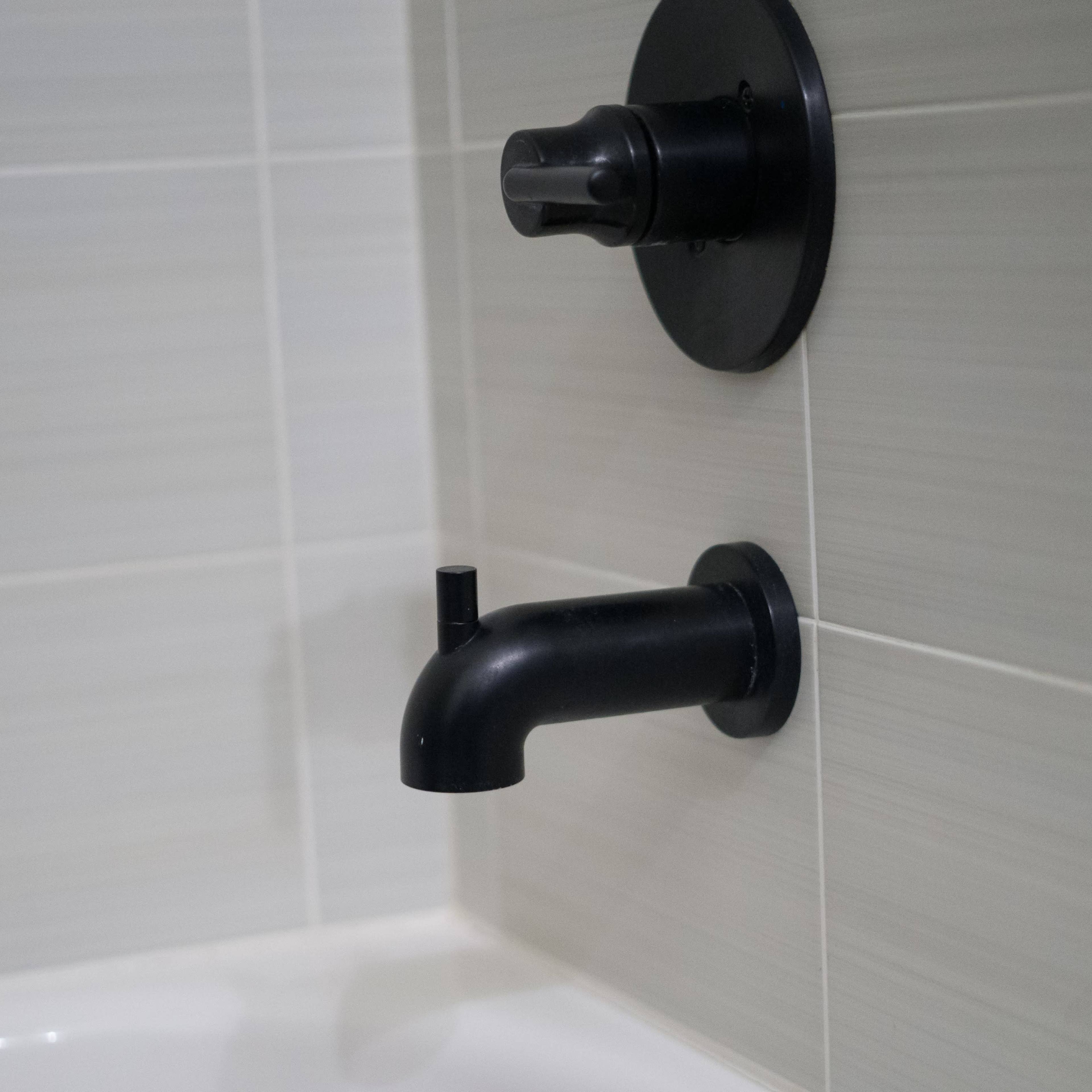 A black faucet and control knob mounted on a light-colored tiled wall in a bathroom.