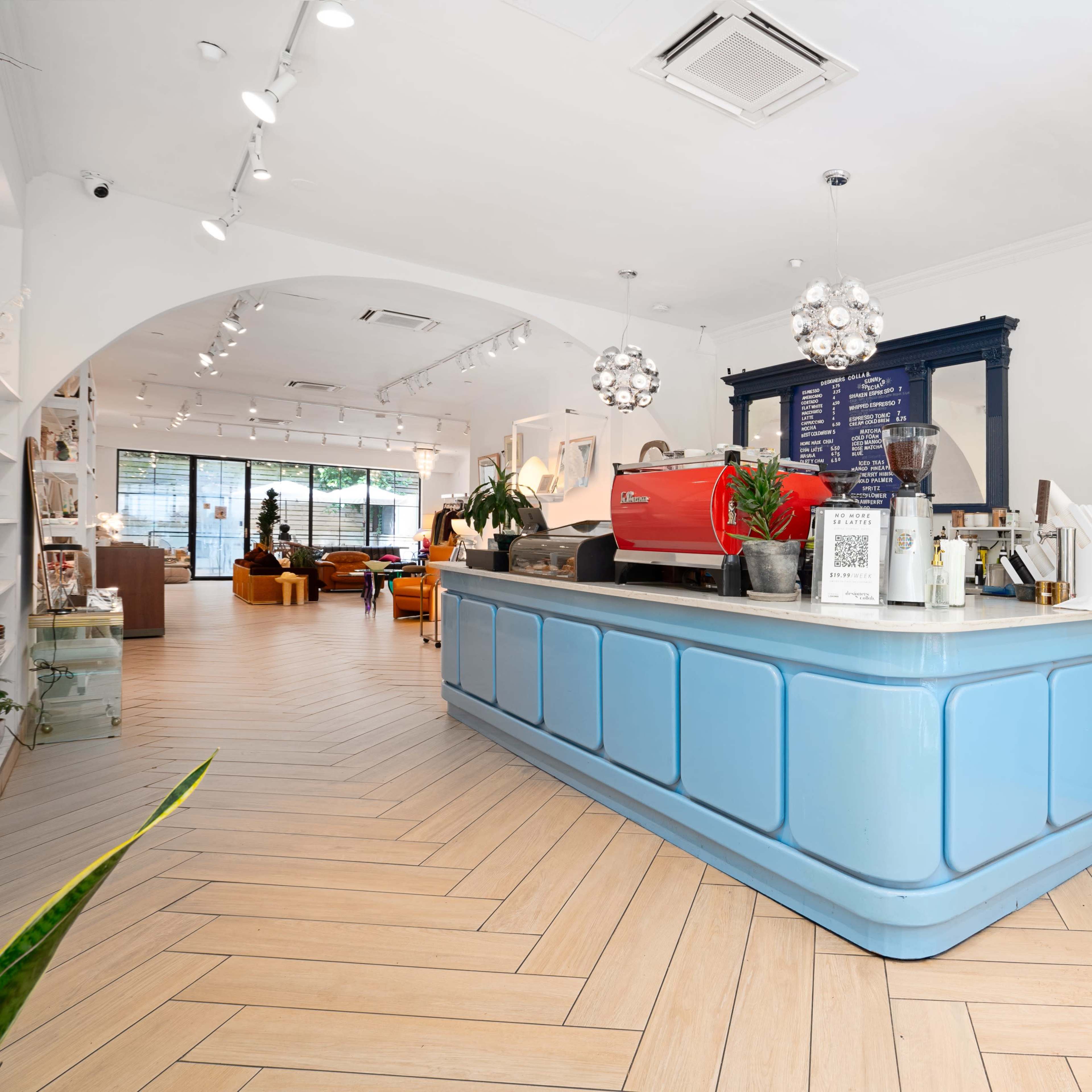 A bright cafe interior with a light blue counter, stylish lighting fixtures, and plants, featuring a coffee machine and a menu on the wall.