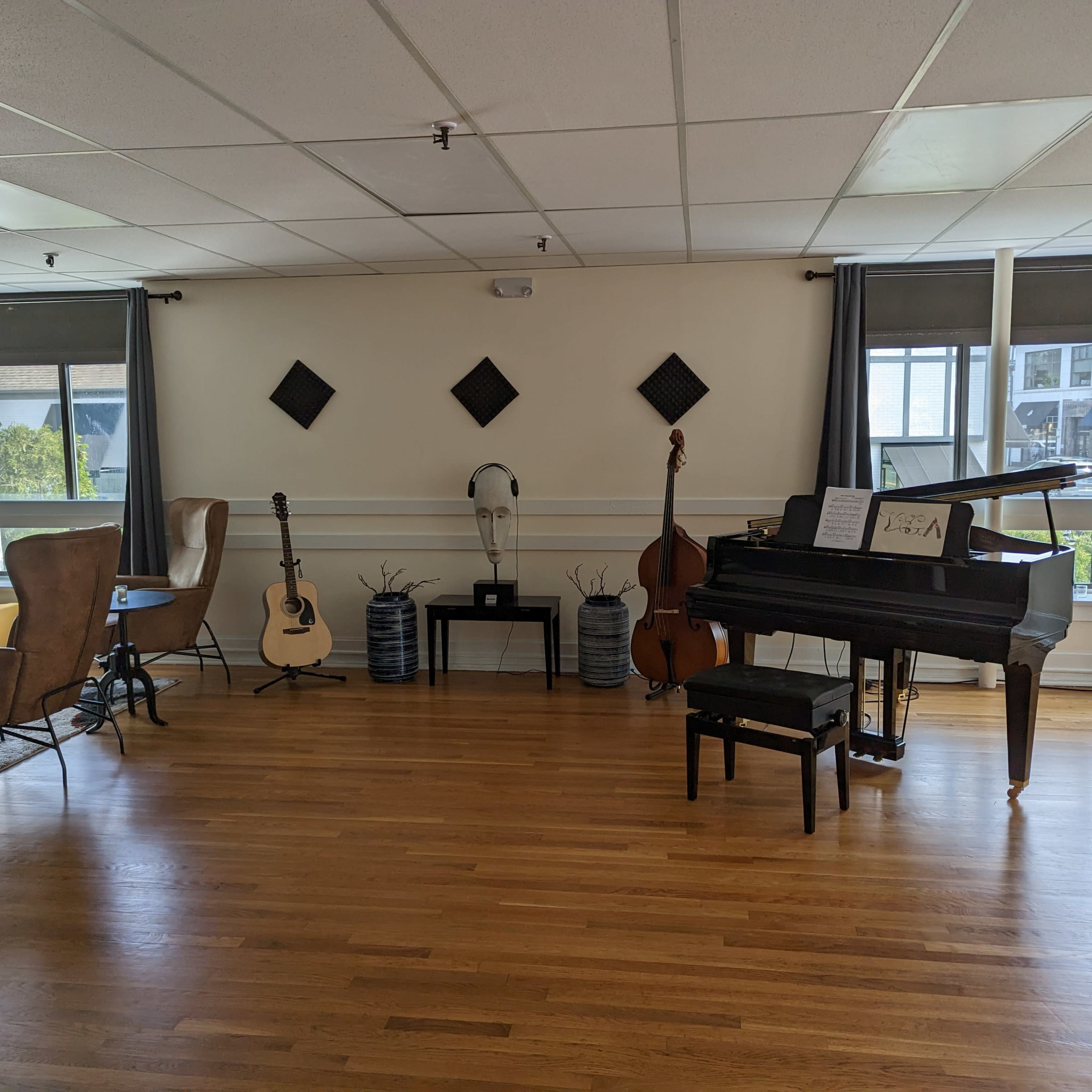 The room features a grand piano, a double bass, an acoustic guitar, and seating areas with lamps, all arranged along wooden flooring under natural light.