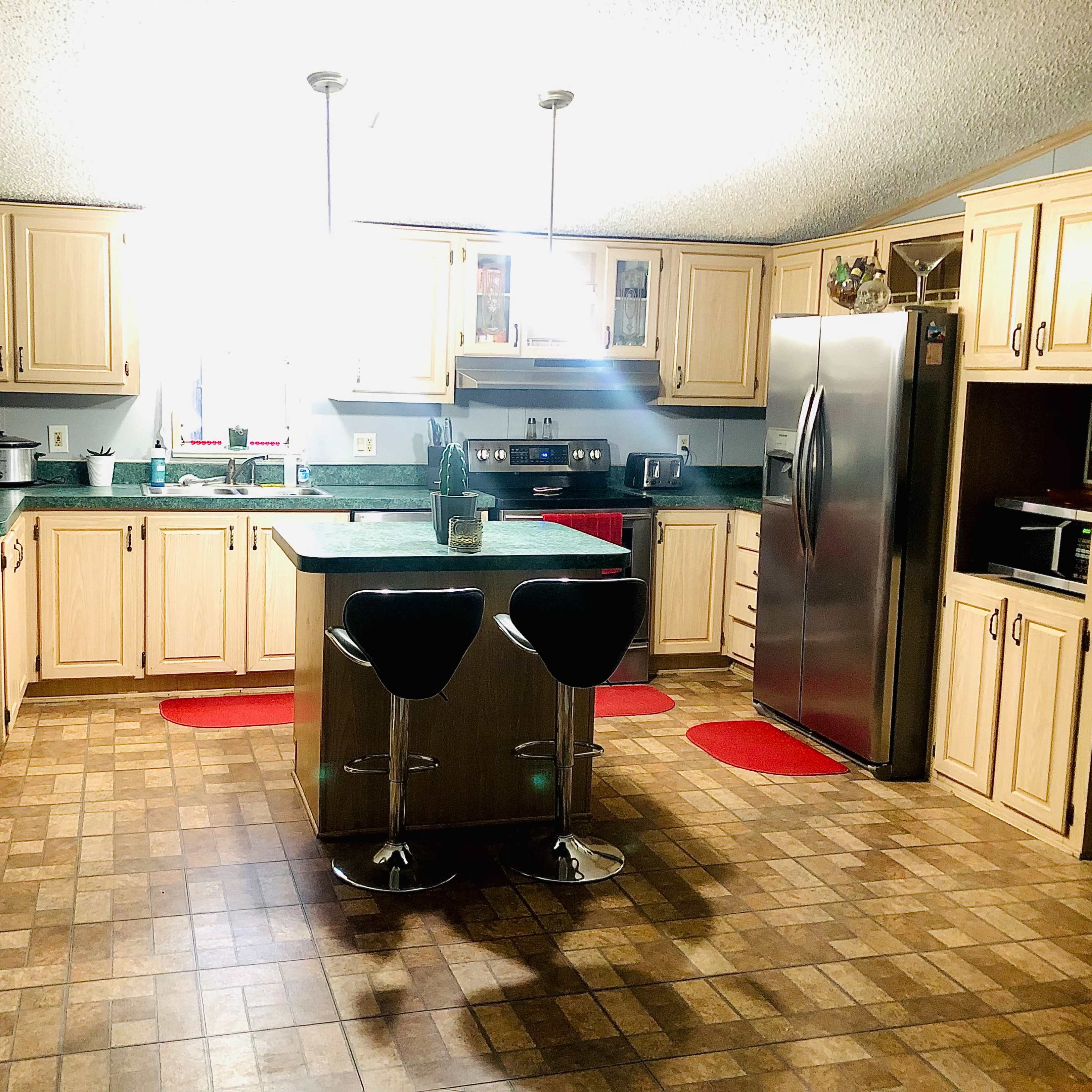 A tidy kitchen with light wood cabinets, a green countertop, stainless steel appliances, and two black bar stools positioned at a central island.