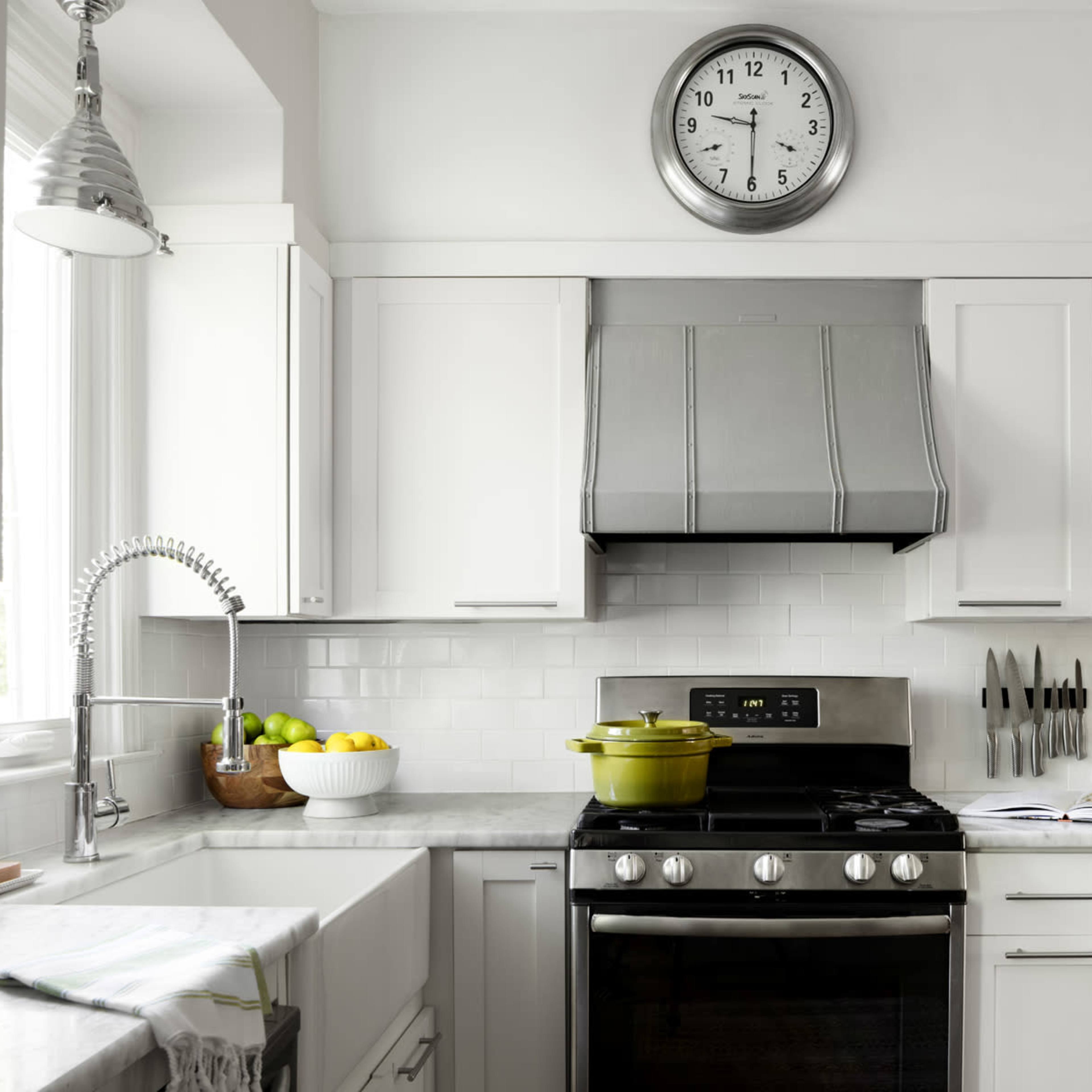 A modern kitchen features white cabinets, a stainless steel range hood, a clock, and a green pot on the stovetop.