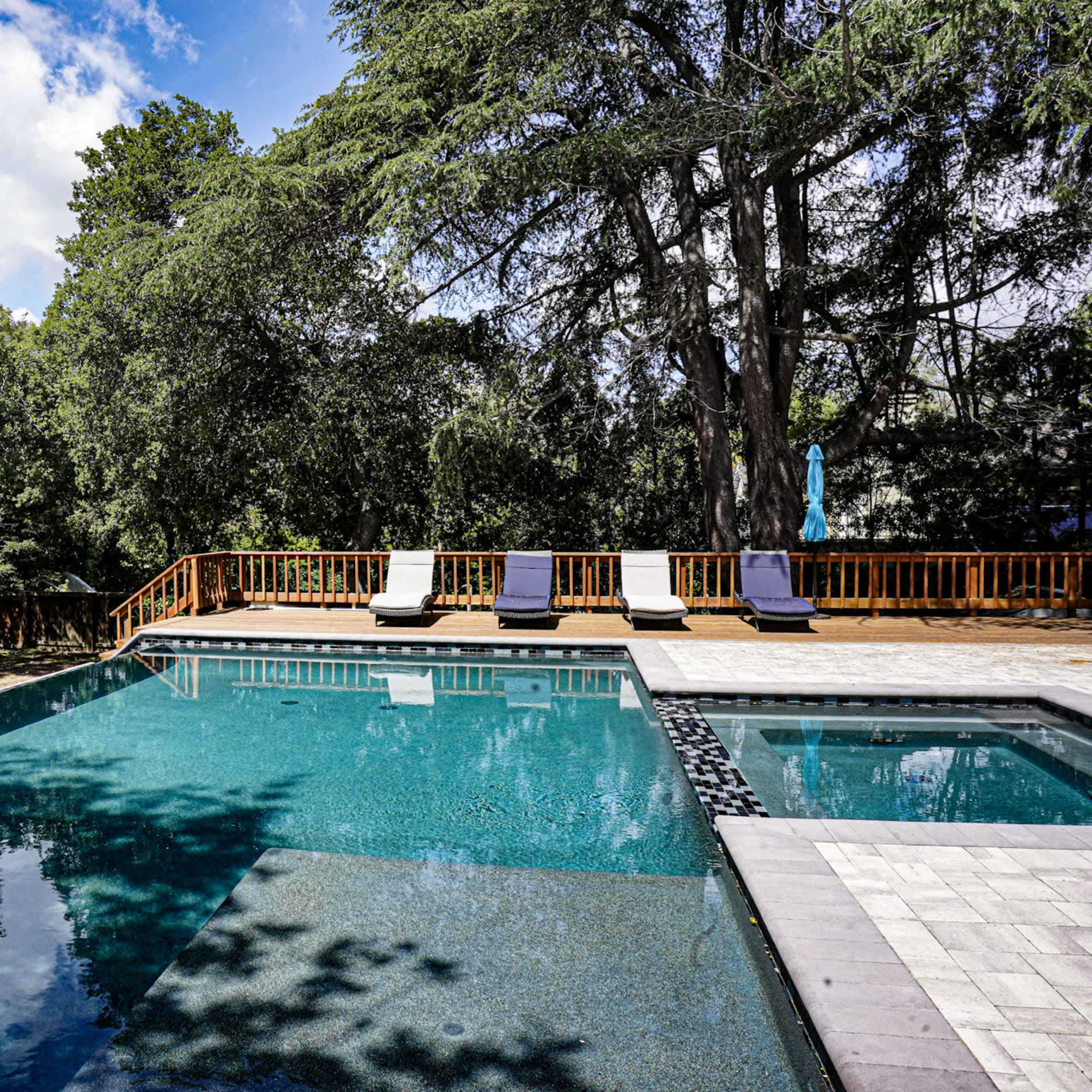 A swimming pool is set in a landscaped area surrounded by trees, with lounge chairs positioned on a wooden deck nearby.