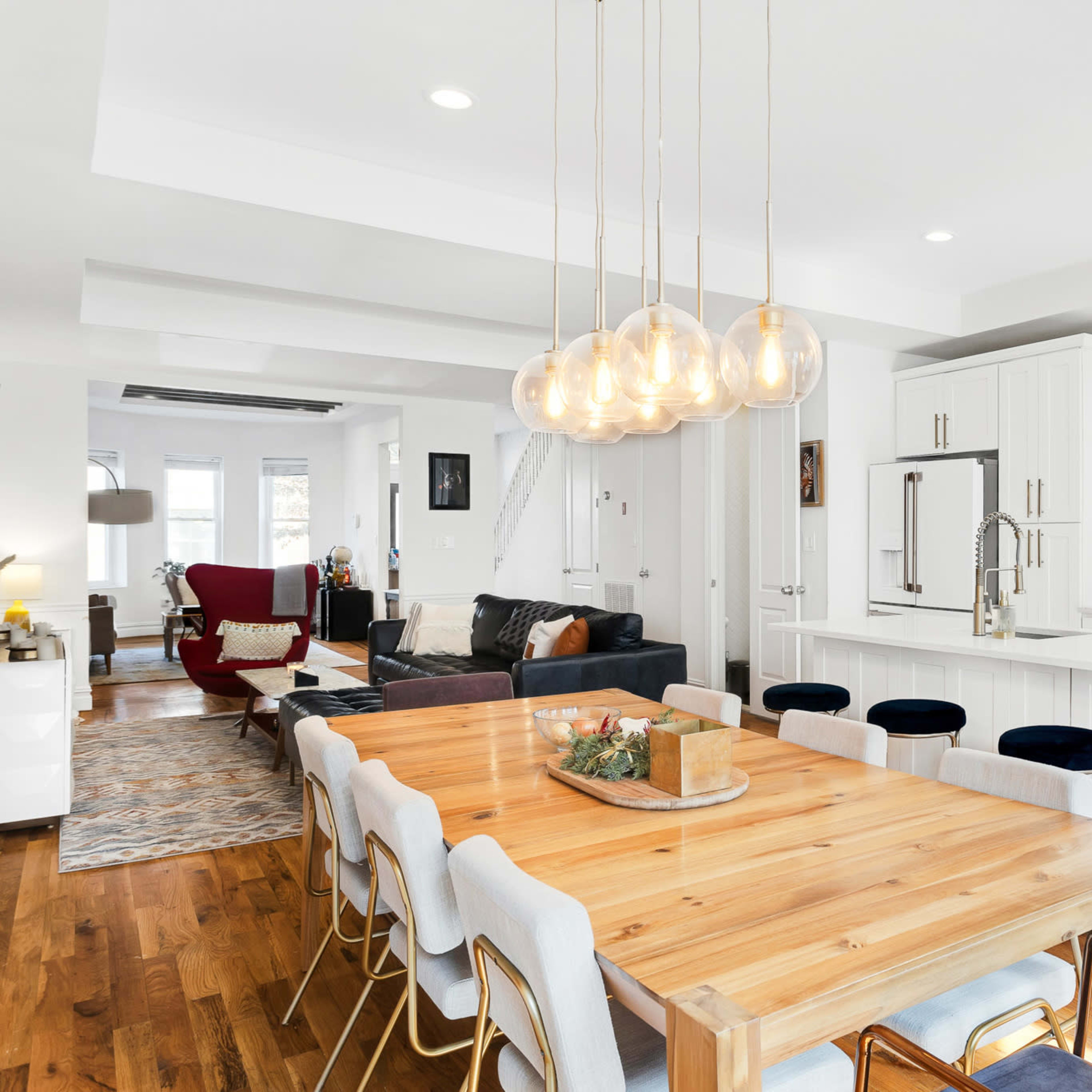 A modern kitchen and dining area features a long wooden table surrounded by upholstered chairs, with pendant lights hanging above and a spacious living area visible in the background.