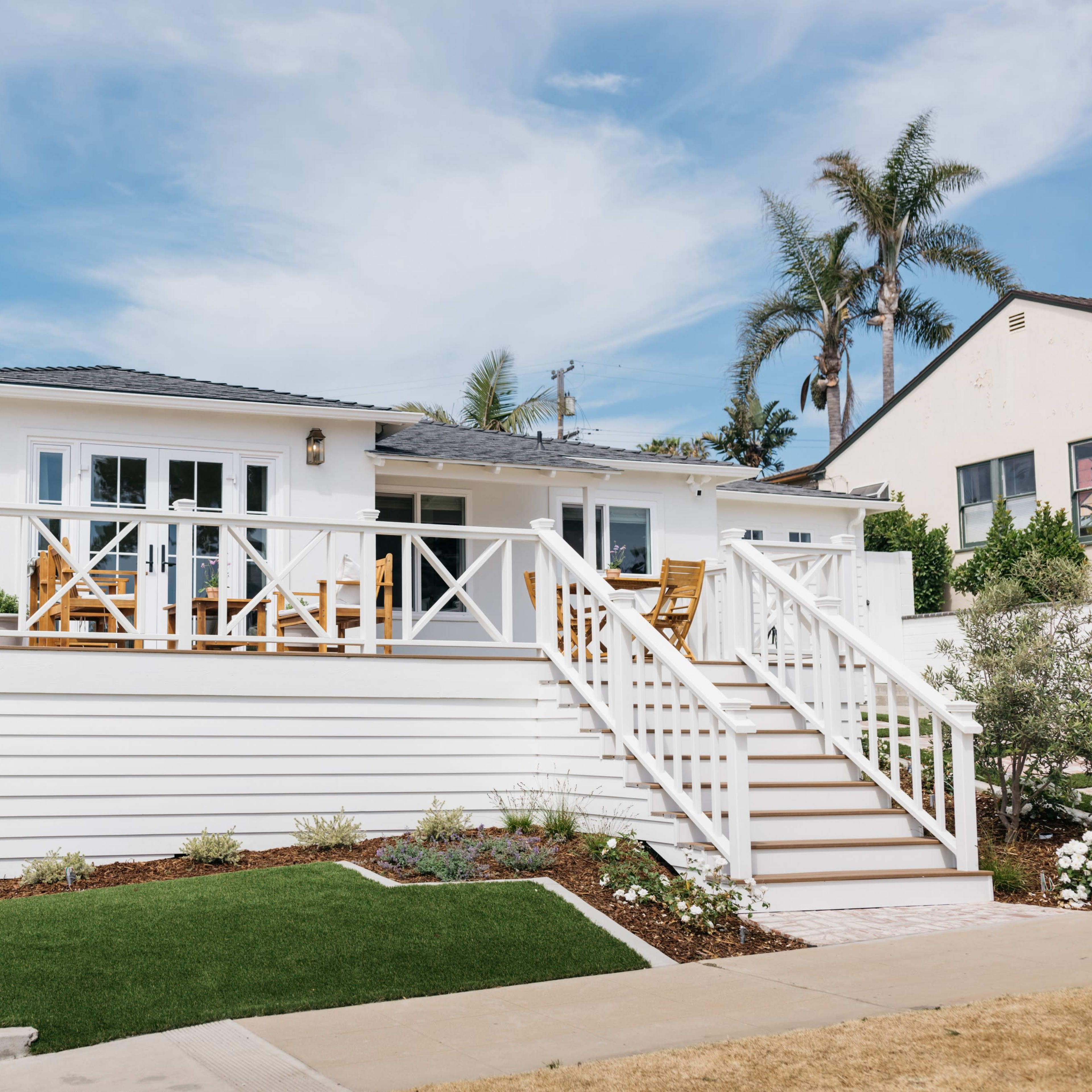 A single-story white house with a porch and staircase is situated on a corner lot, surrounded by landscaped greenery and palm trees.