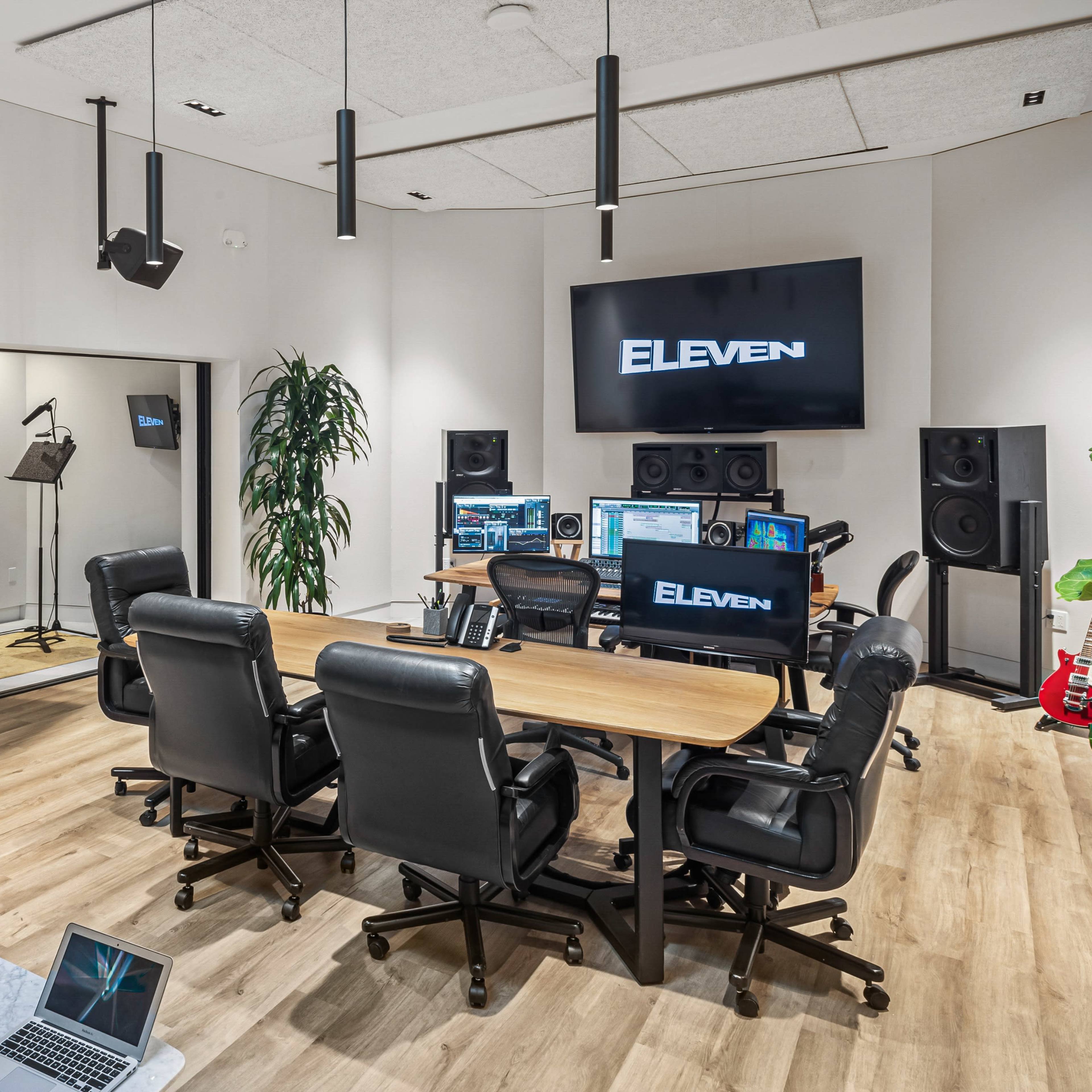 A modern recording studio equipped with a large screen displaying "ELEven," multiple computer workstations, audio monitors, and a small seating area.