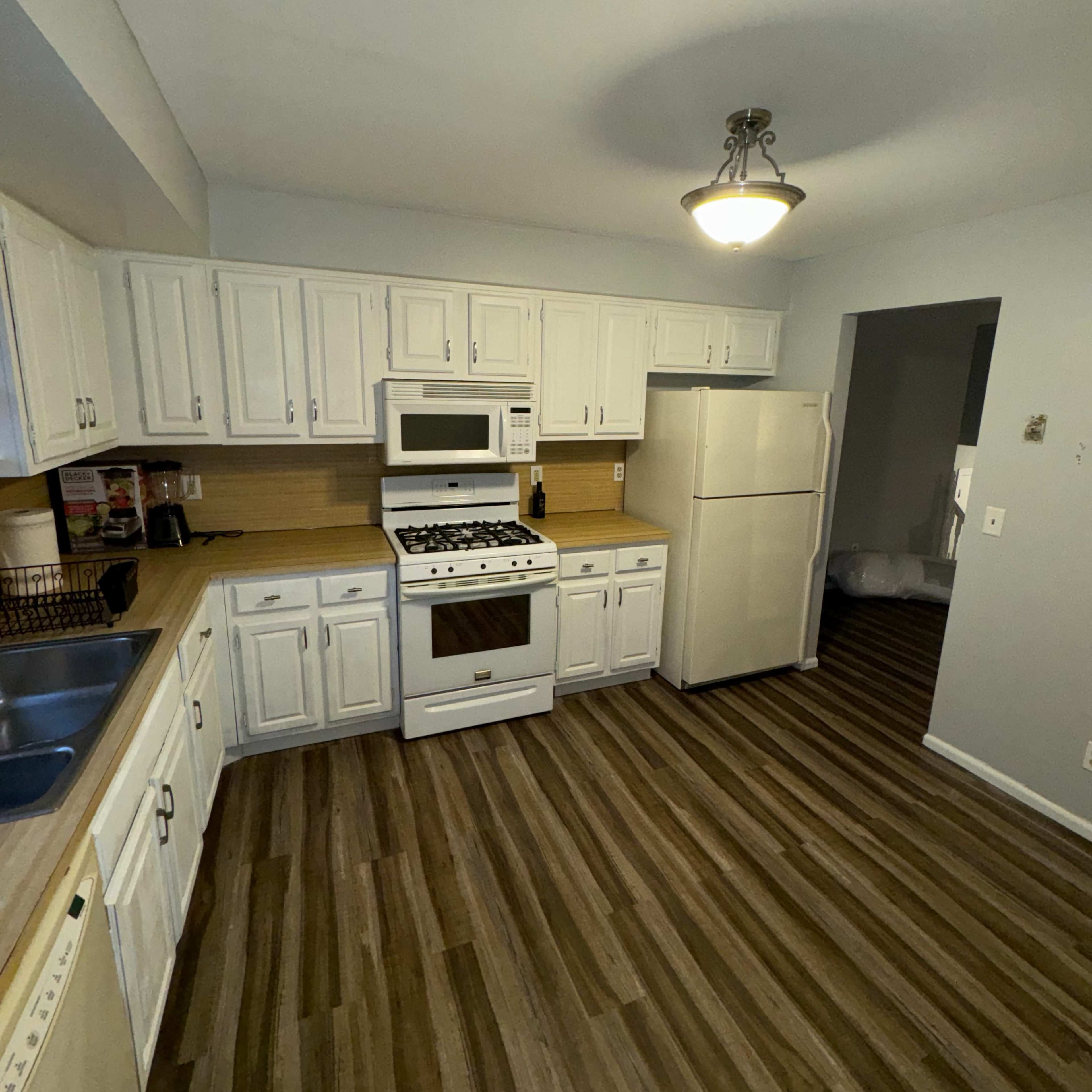 A kitchen with white cabinetry, a gas stove, microwave, and a refrigerator, all set against a wood-patterned floor.