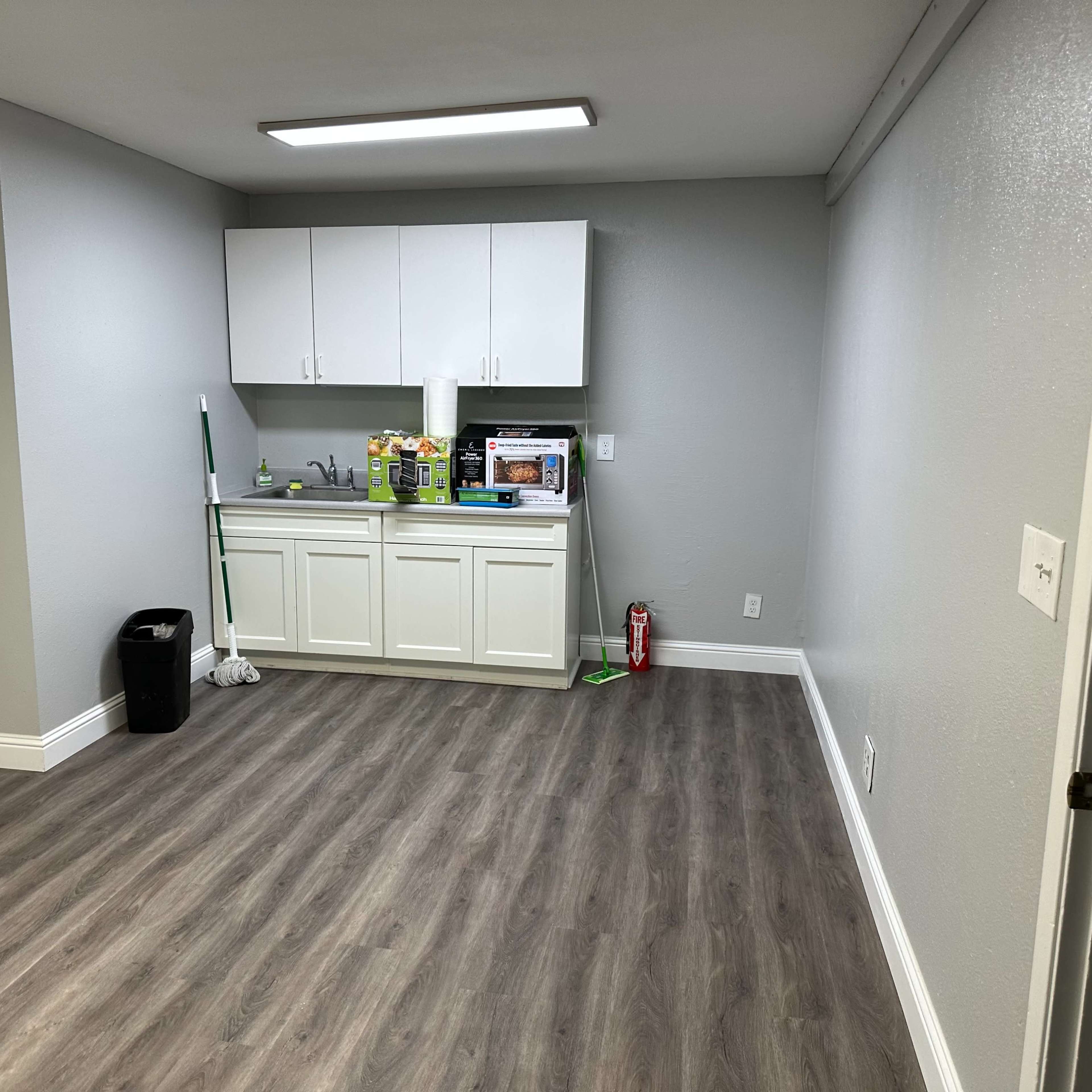 A clean, empty room with a kitchenette featuring white cabinets and a countertop, along with a mop, a trash can, and a fire extinguisher.