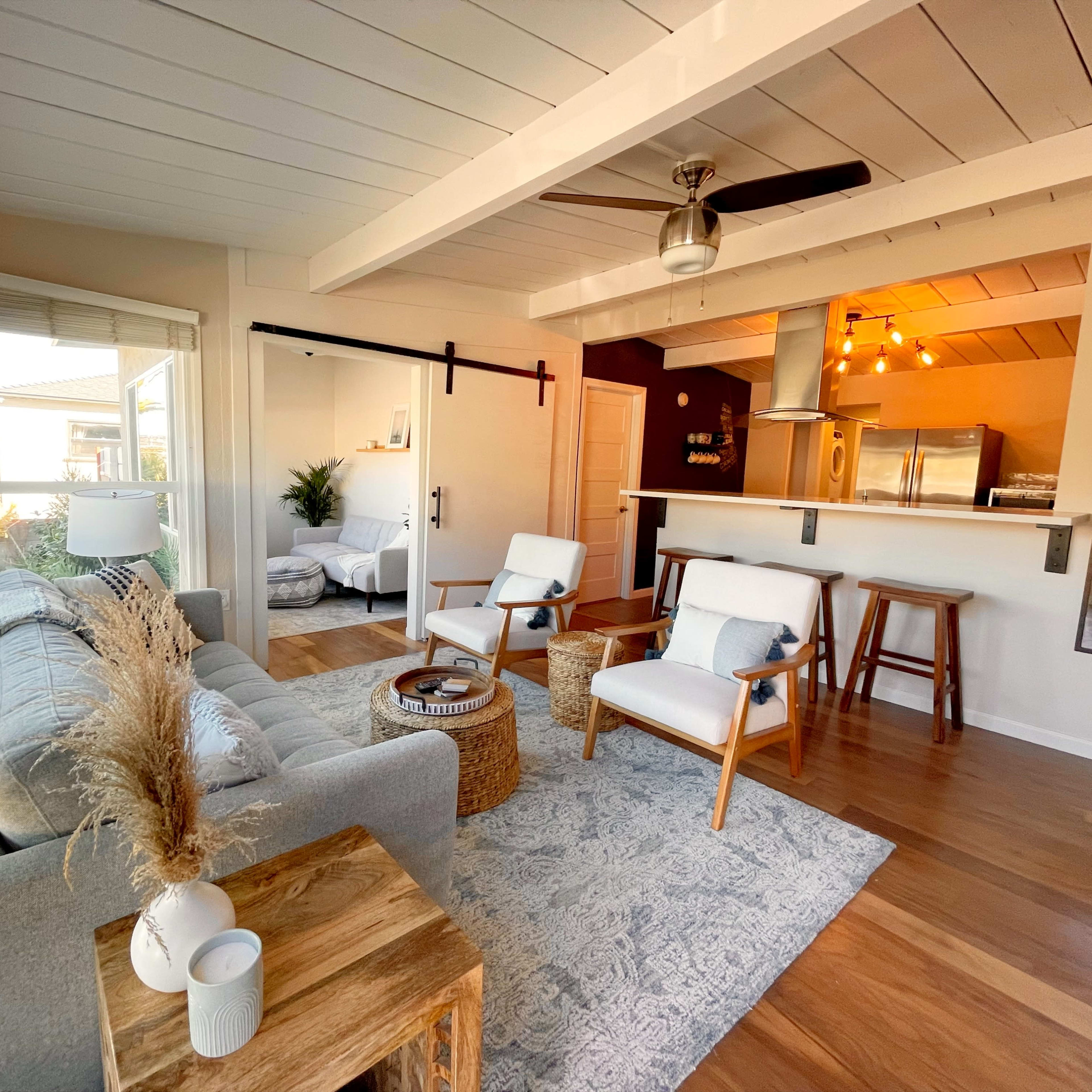 A modern living room with a gray sofa, two white chairs, a wooden coffee table, and an open kitchen in the background, featuring a bar counter and stainless steel appliances.
