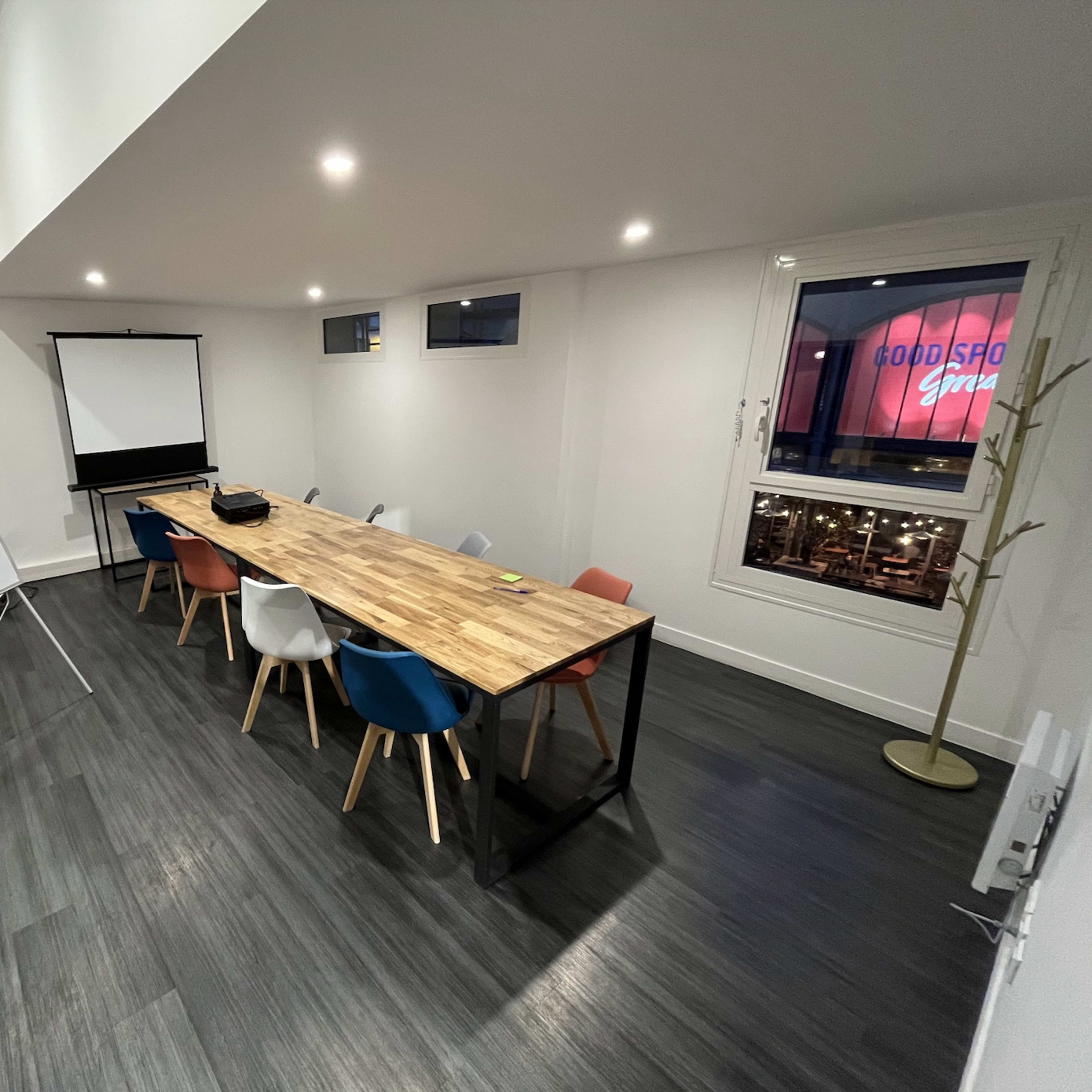 A wooden conference table is surrounded by colorful chairs in a bright, modern meeting room with a projector screen.