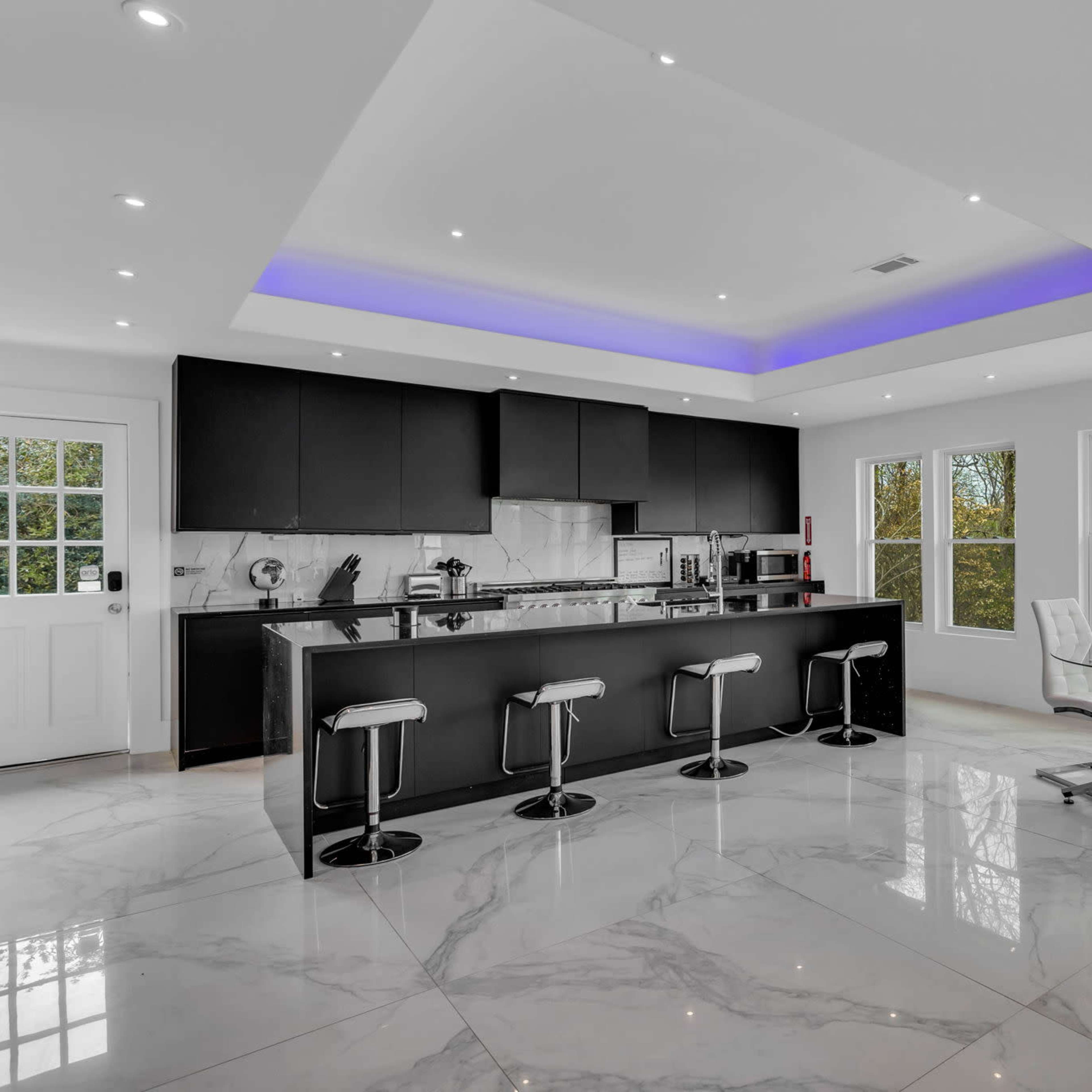 A modern kitchen with a central island, bar stools, and large windows allowing natural light to illuminate the marble floor.