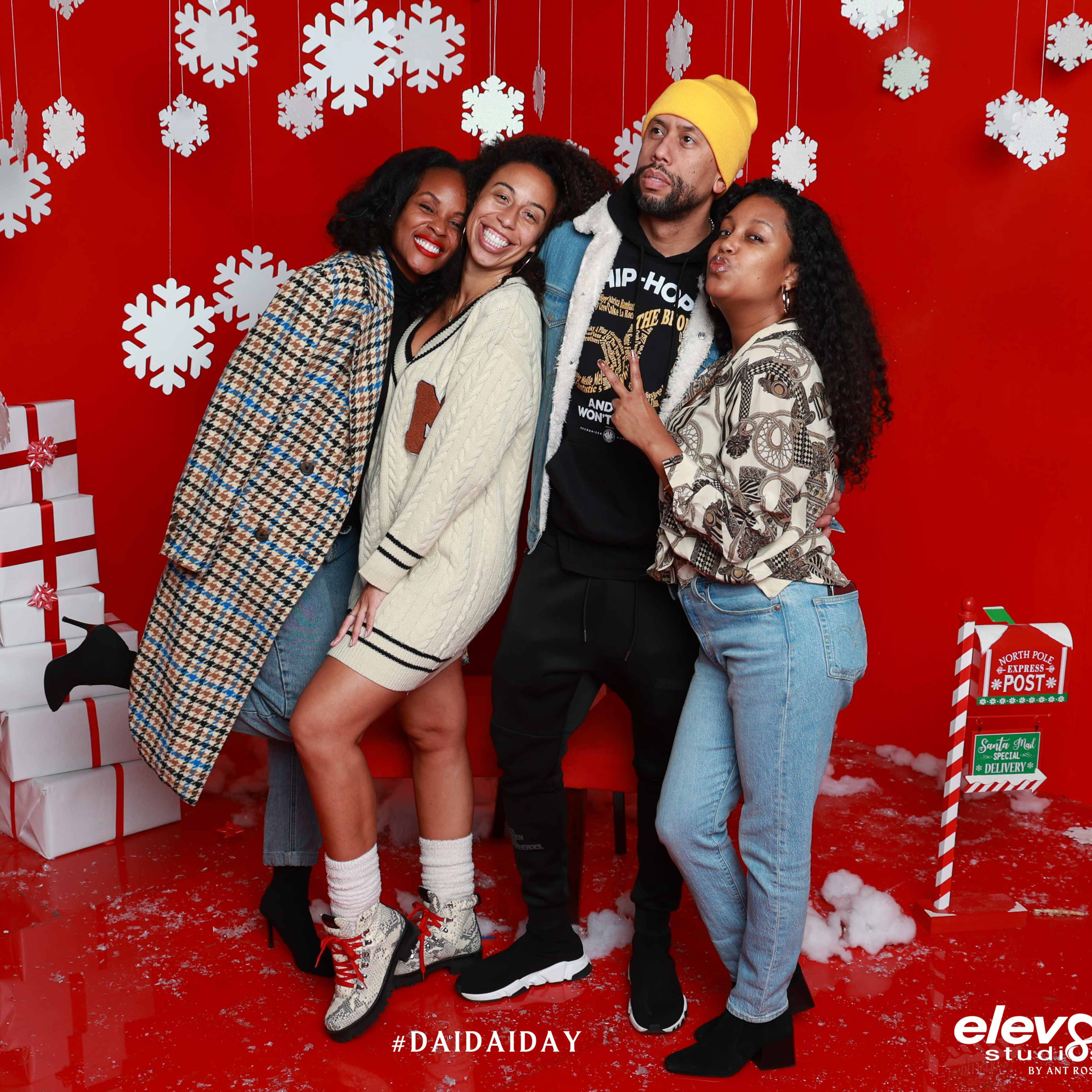 A group of four people poses together in front of a festive red backdrop with Christmas decorations and gifts.