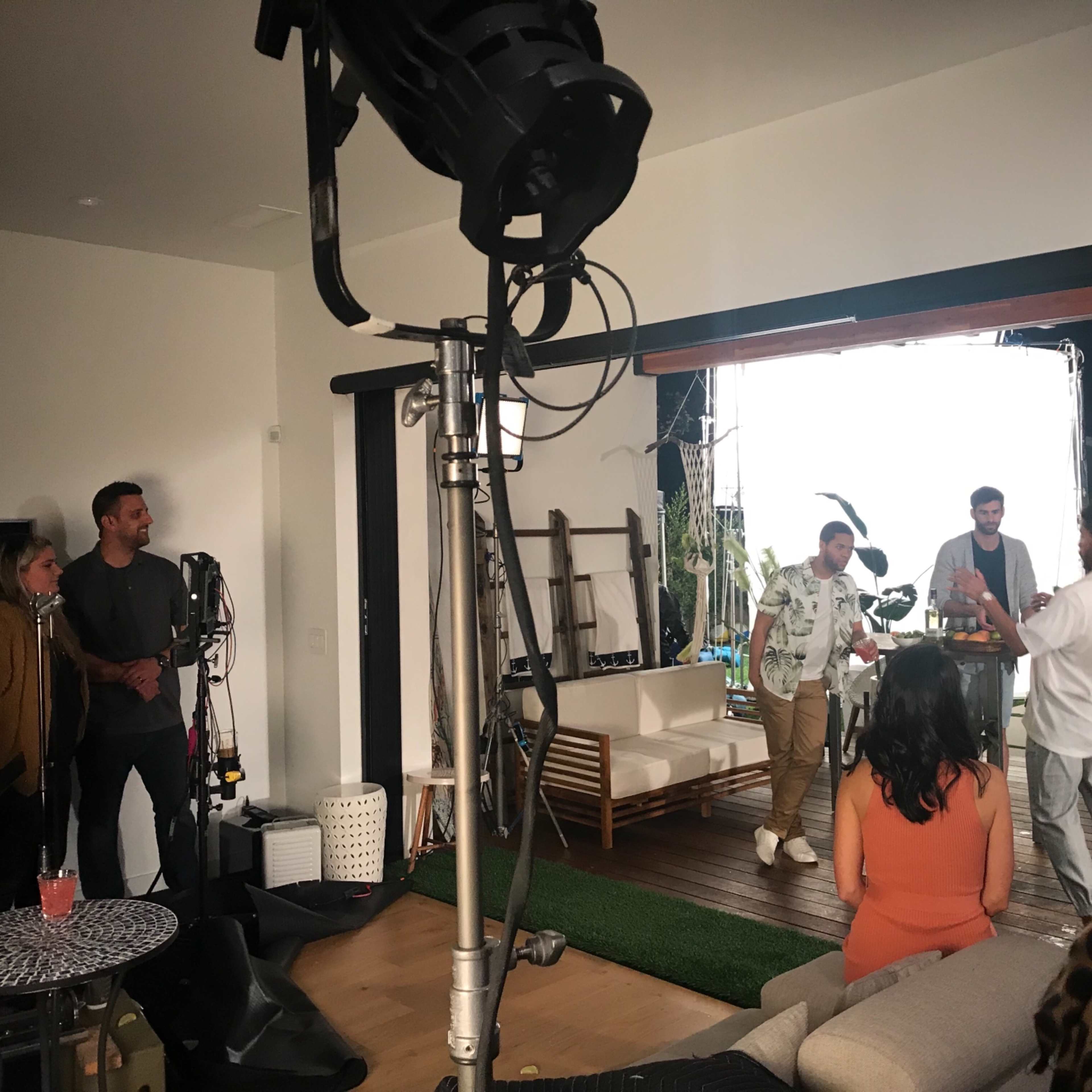 A group of five people is gathered around a table, engaged in conversation, while filming takes place in a modern living room setting.