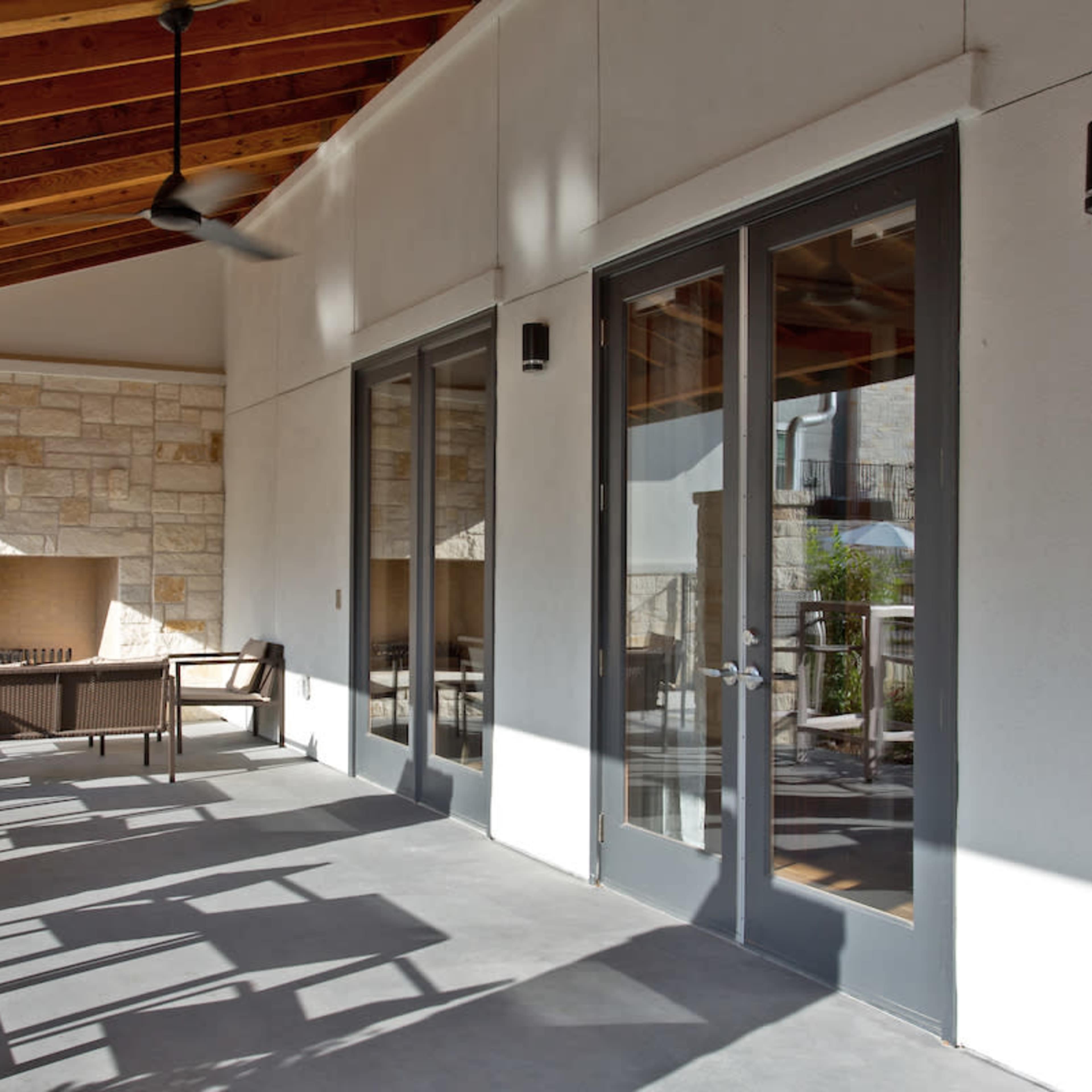 A covered patio area with stone walls, large glass doors, and a seating arrangement beside a fireplace.