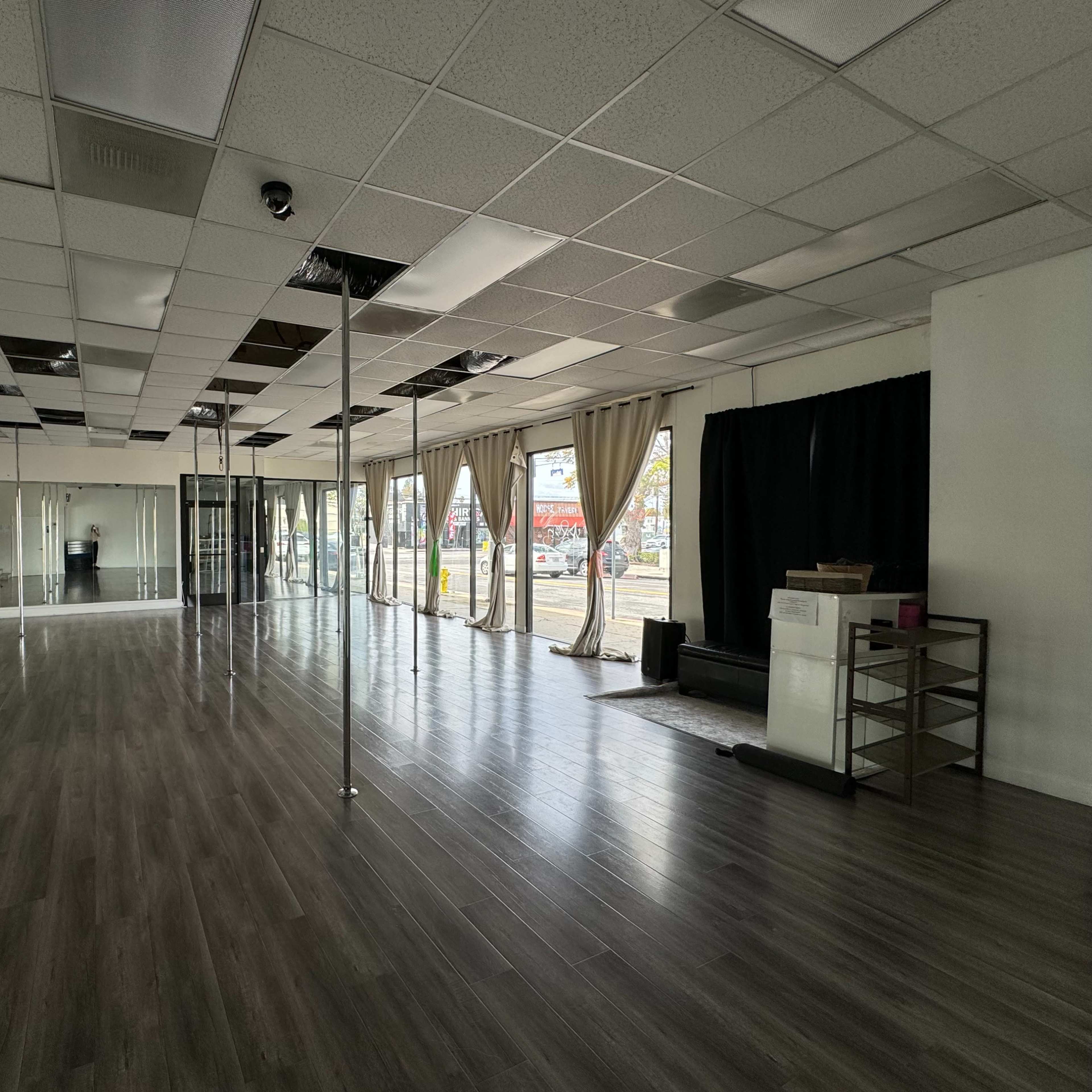 An empty dance studio with several dance poles, a mirrored wall, and large windows revealing an outdoor view.