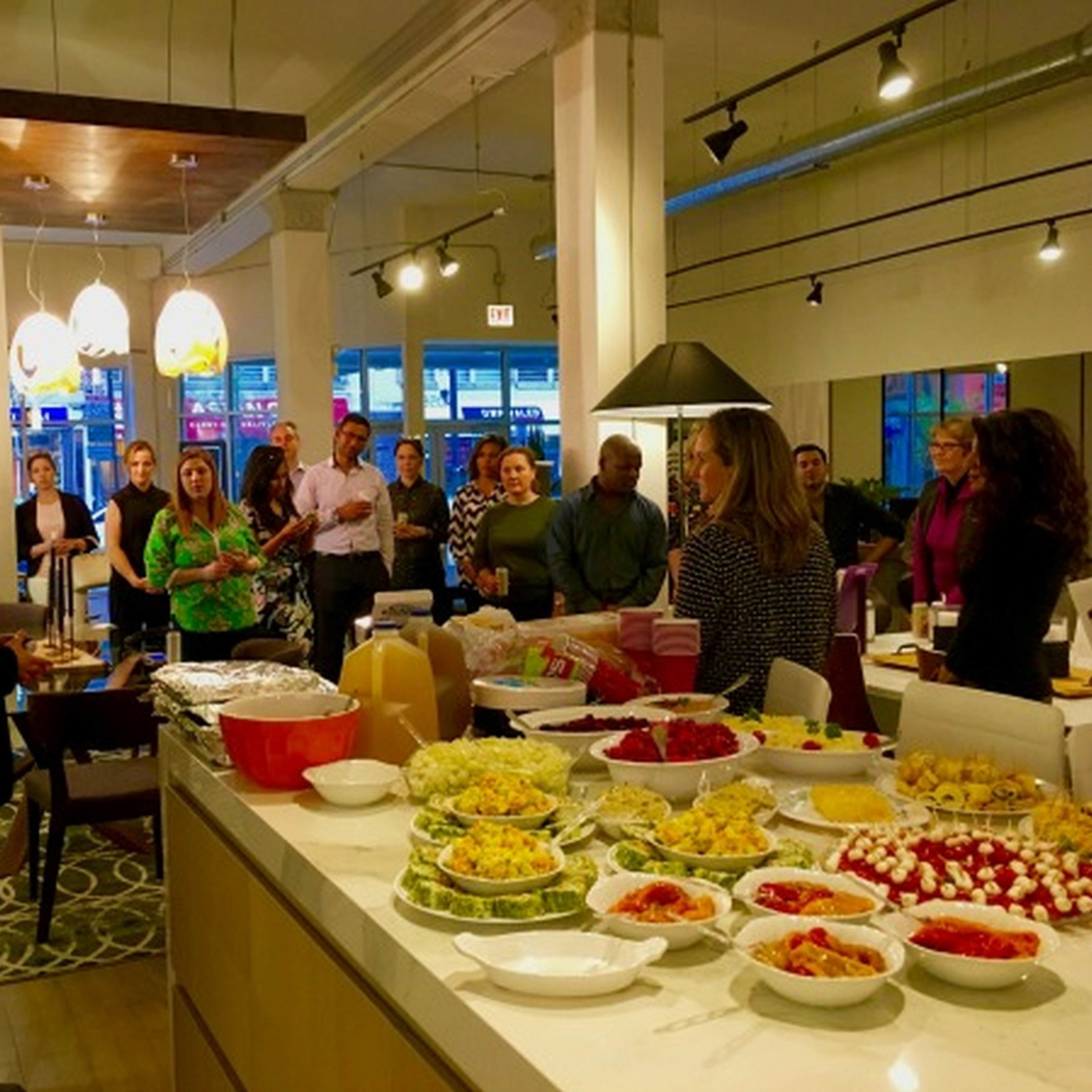 A group of people gathers around a man speaking in a modern kitchen space filled with various dishes on a countertop.