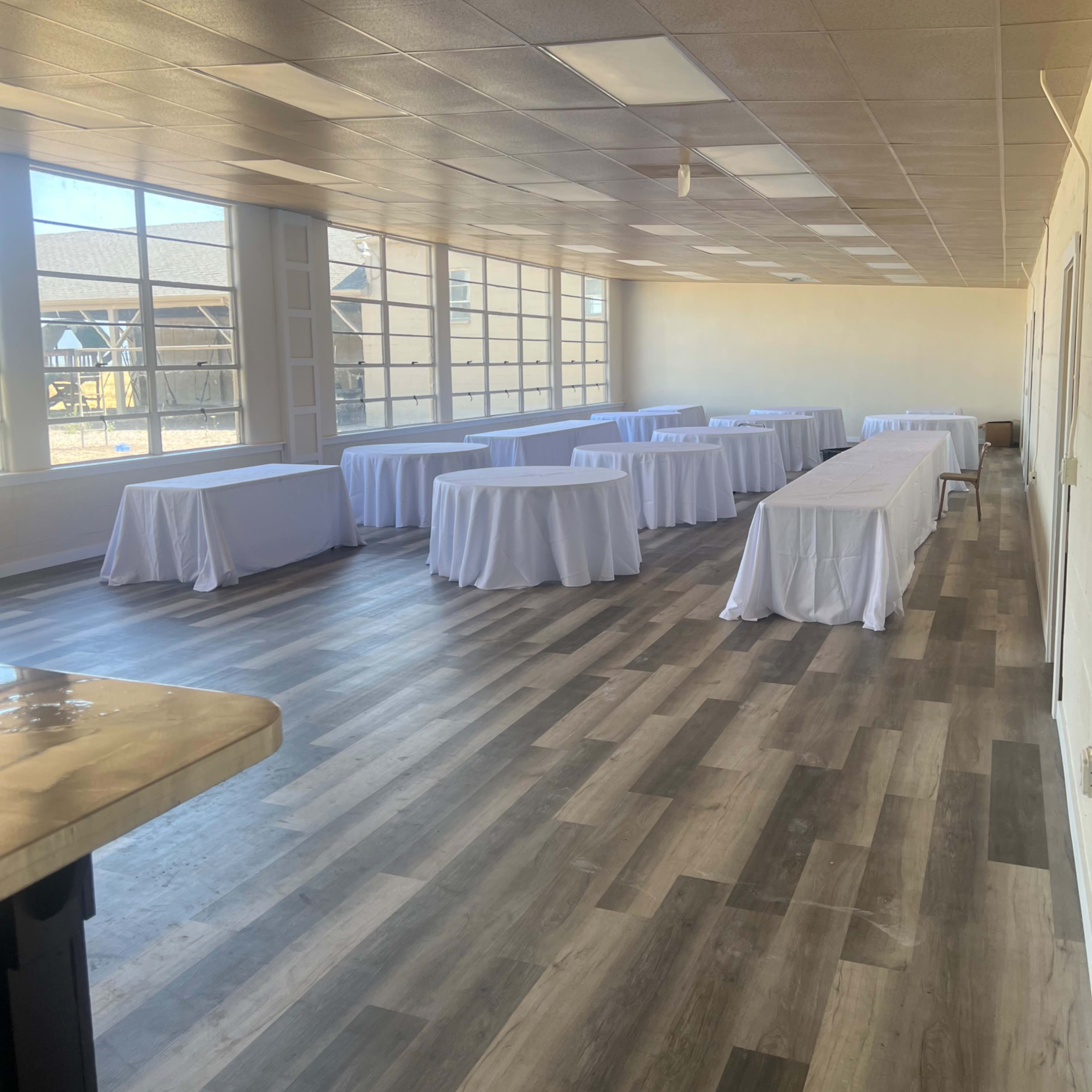 A spacious room with multiple tables covered in white tablecloths, illuminated by natural light from large windows.