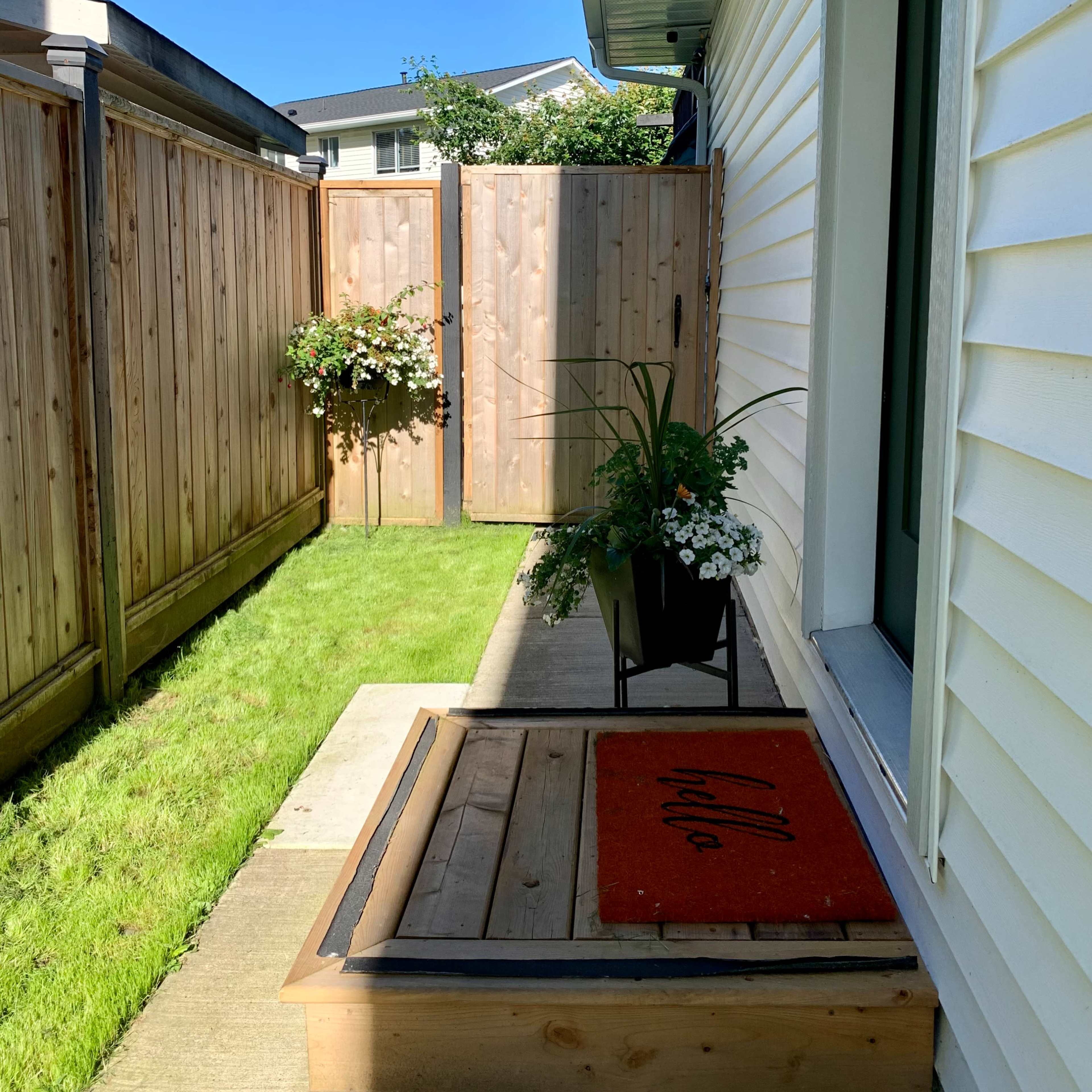 A welcome mat is positioned in front of a small entrance beside a garden with wooden fences and potted plants.