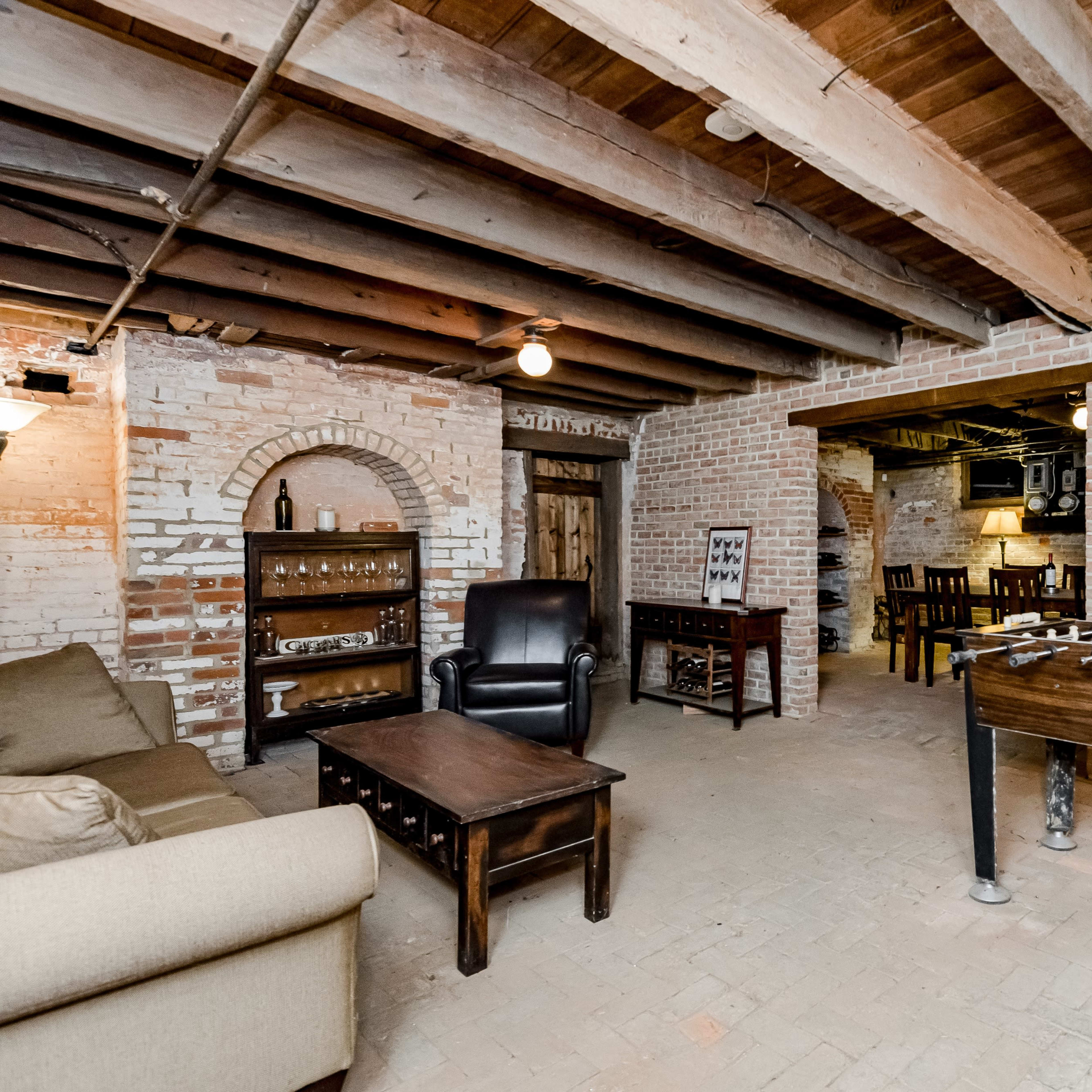 A cozy basement lounge area with exposed brick walls, wooden beams, a sofa, a coffee table, a foosball table, and a small dining area in the background.