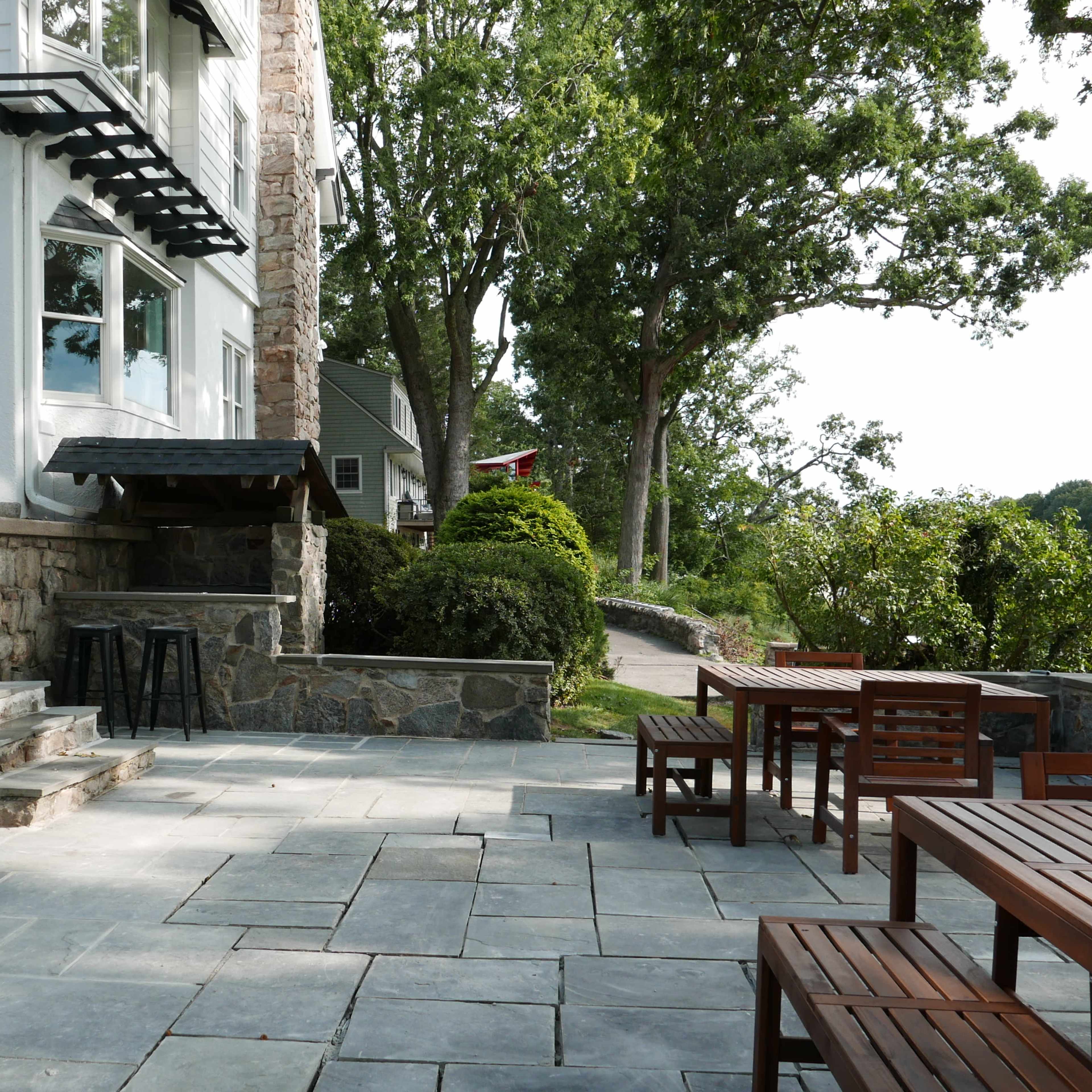 A stone patio with wooden tables and chairs is situated next to a house, surrounded by greenery and leading toward a pathway.