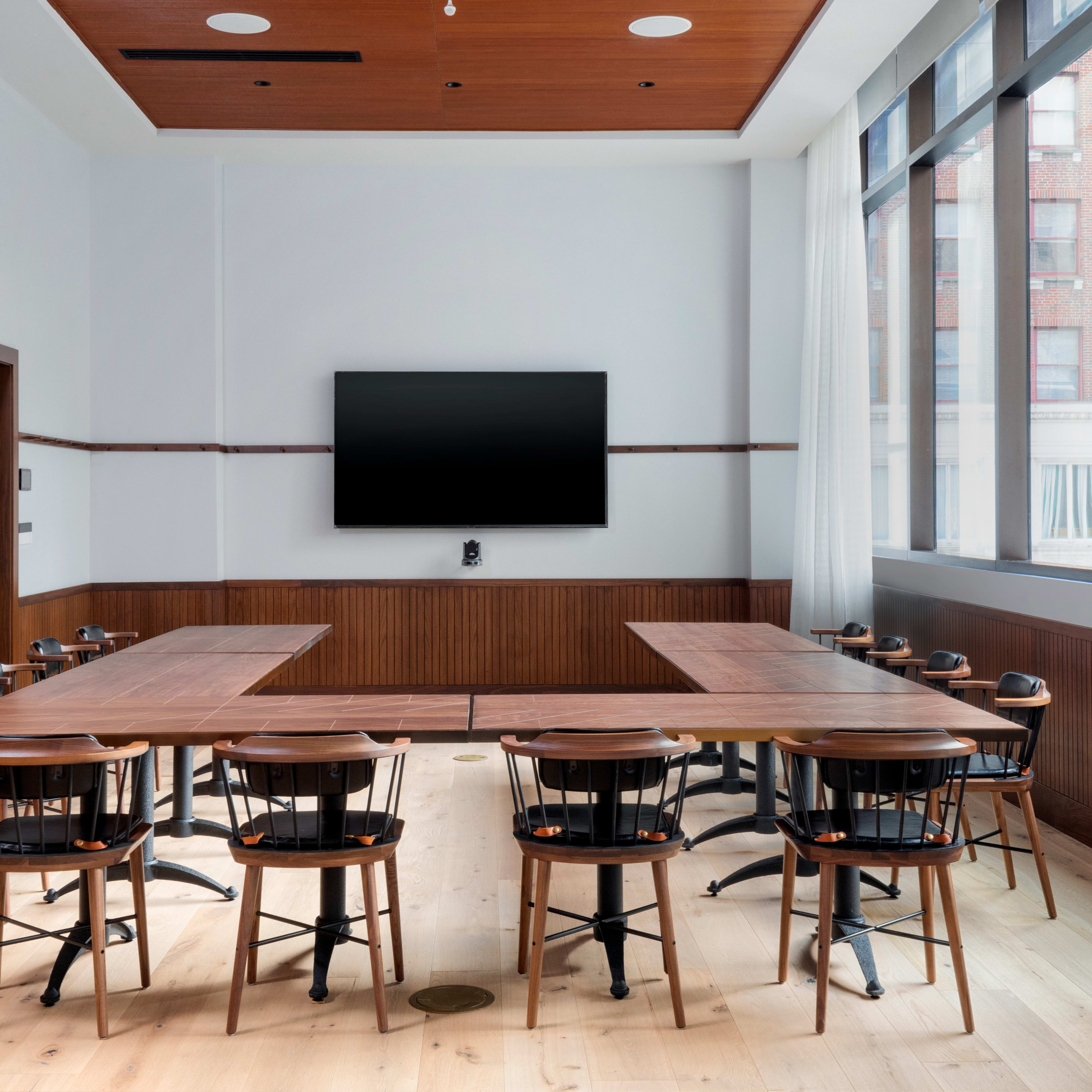 A modern conference room features a lengthy wooden table surrounded by black chairs, with a large screen mounted on the wall and windows allowing natural light.