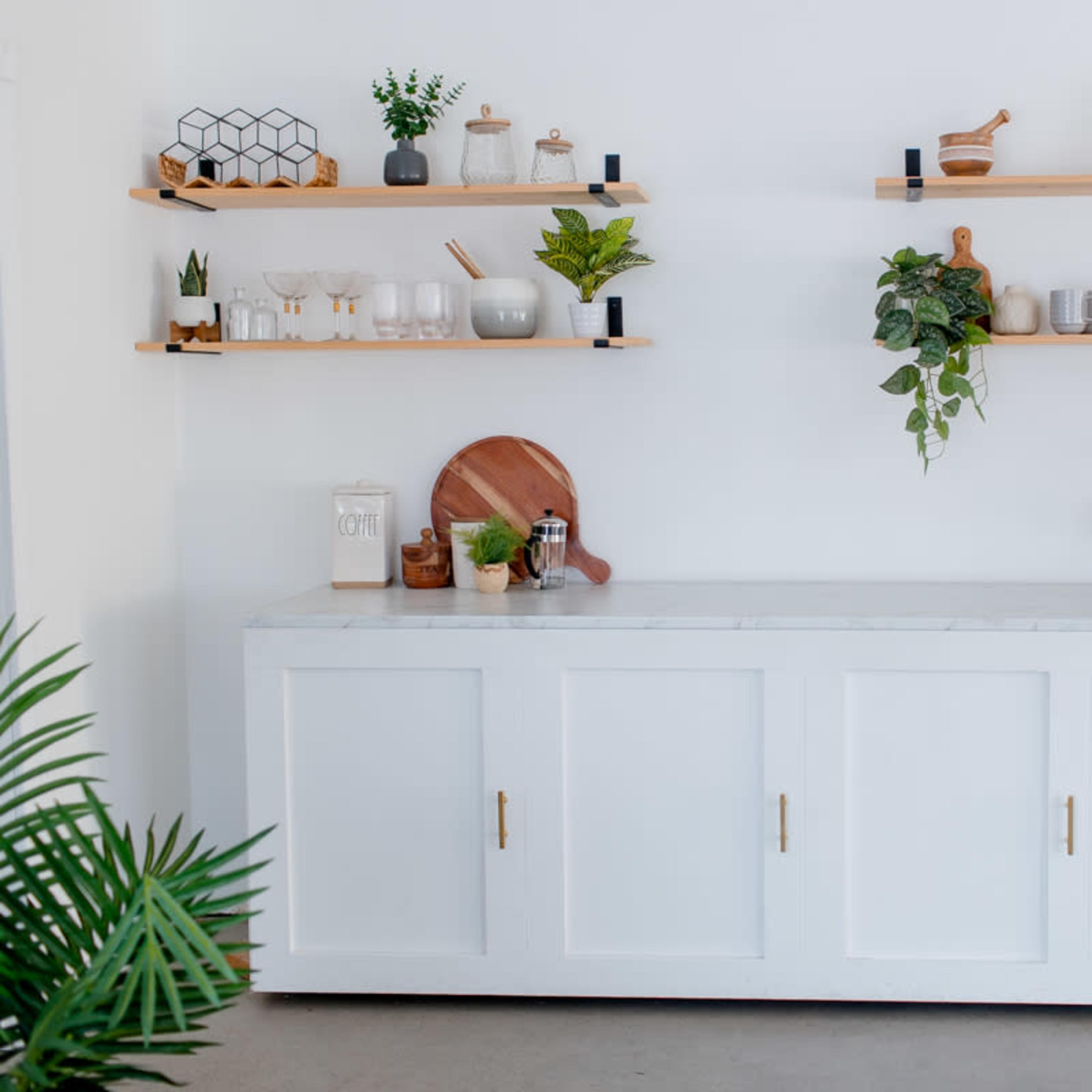A clean white cabinet with three doors beneath wooden shelves that hold various kitchenware and decorative plants.