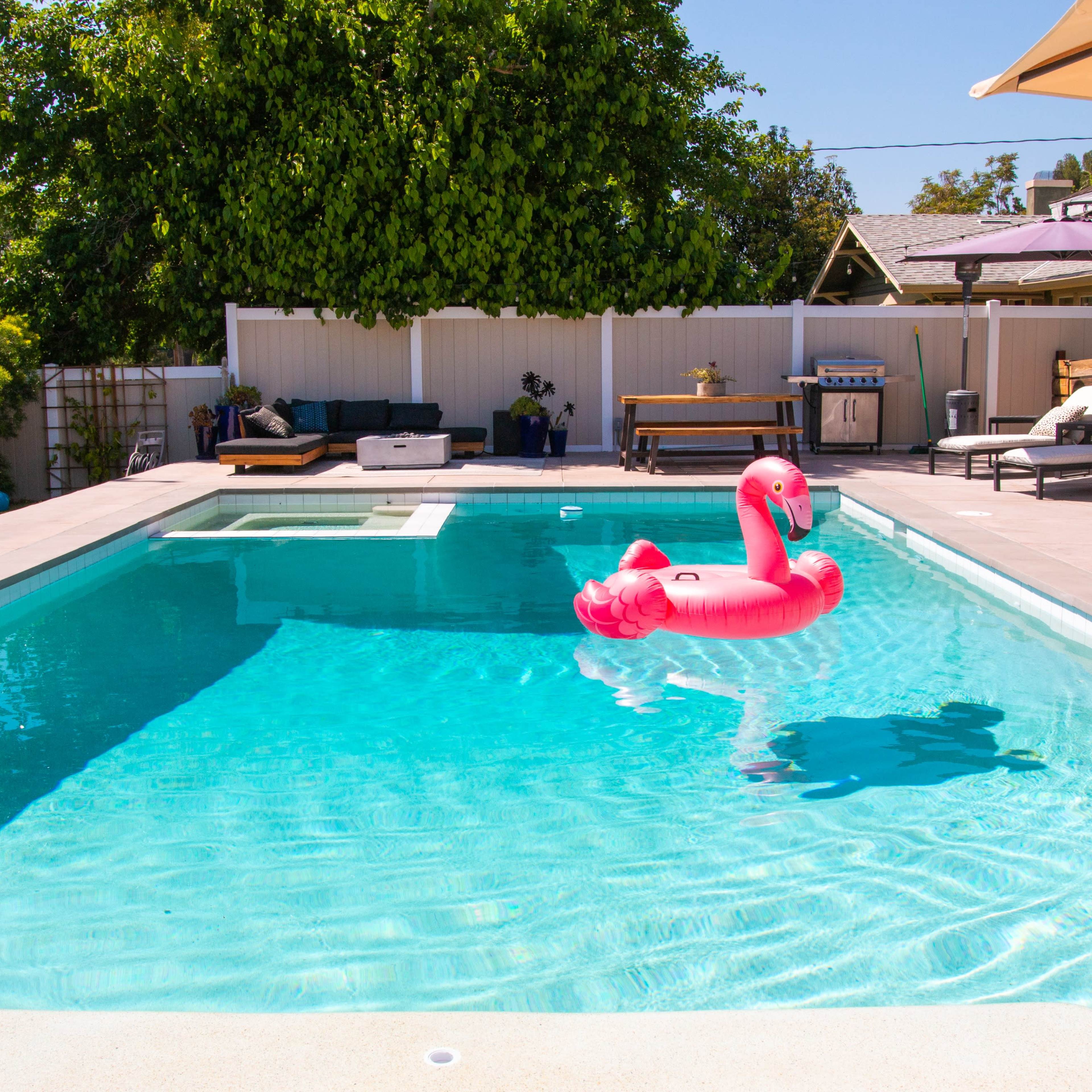 A pink flamingo float sits in a clear swimming pool surrounded by lounge chairs and a shaded seating area.