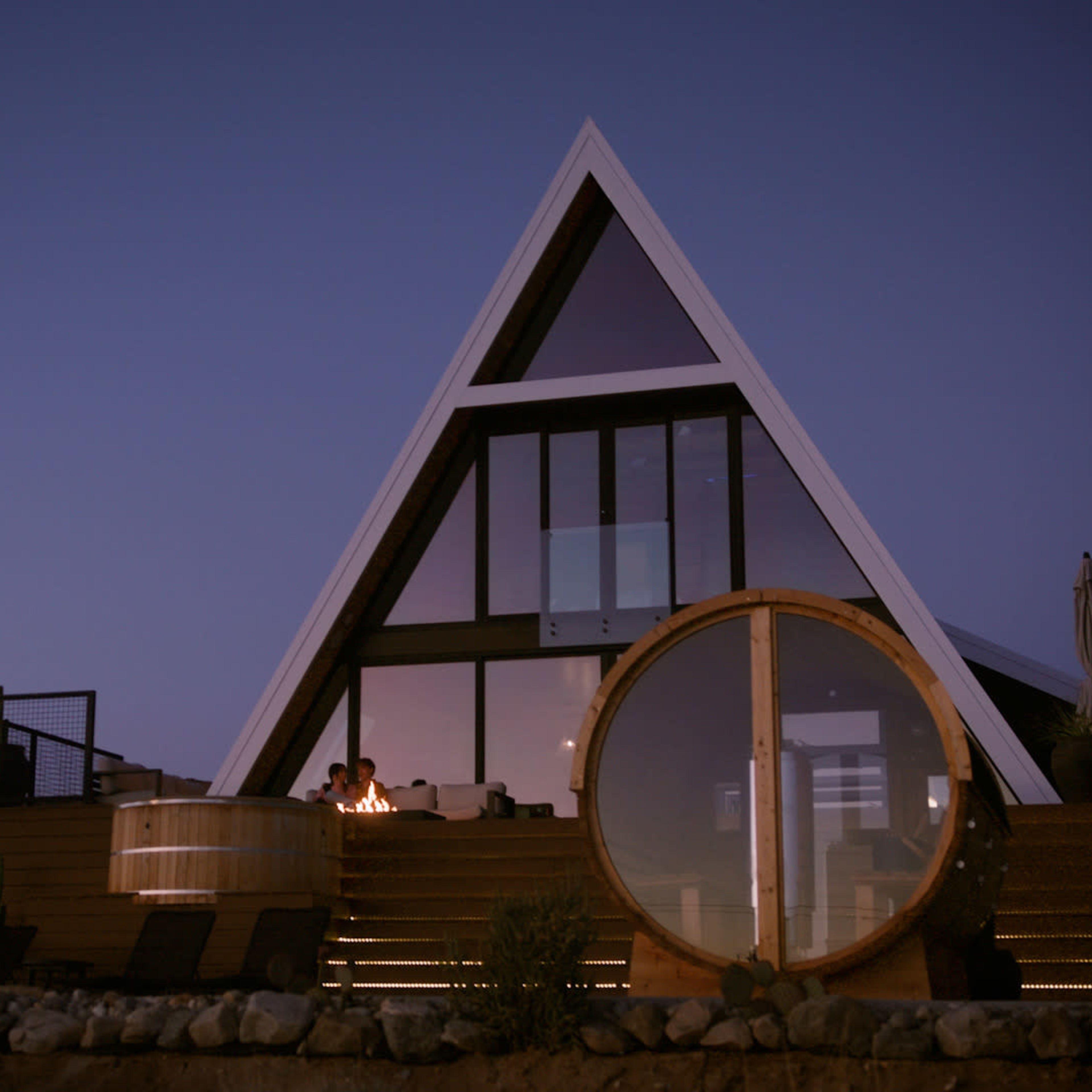 A modern A-frame house with large windows and a circular hot tub is set against a twilight sky.