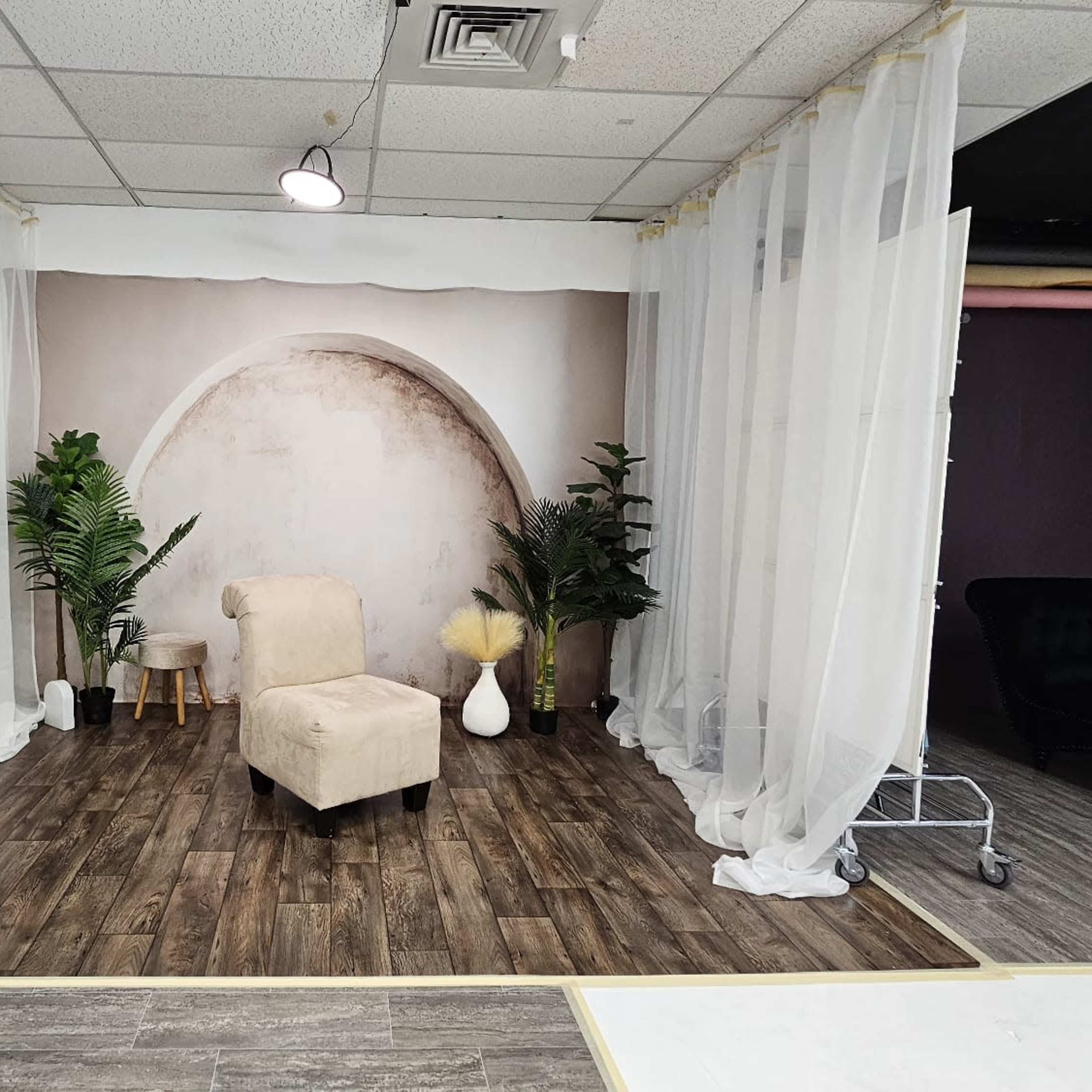 A studio setup with a light-colored armchair facing a backdrop of an arched design, surrounded by potted plants and soft drapery.