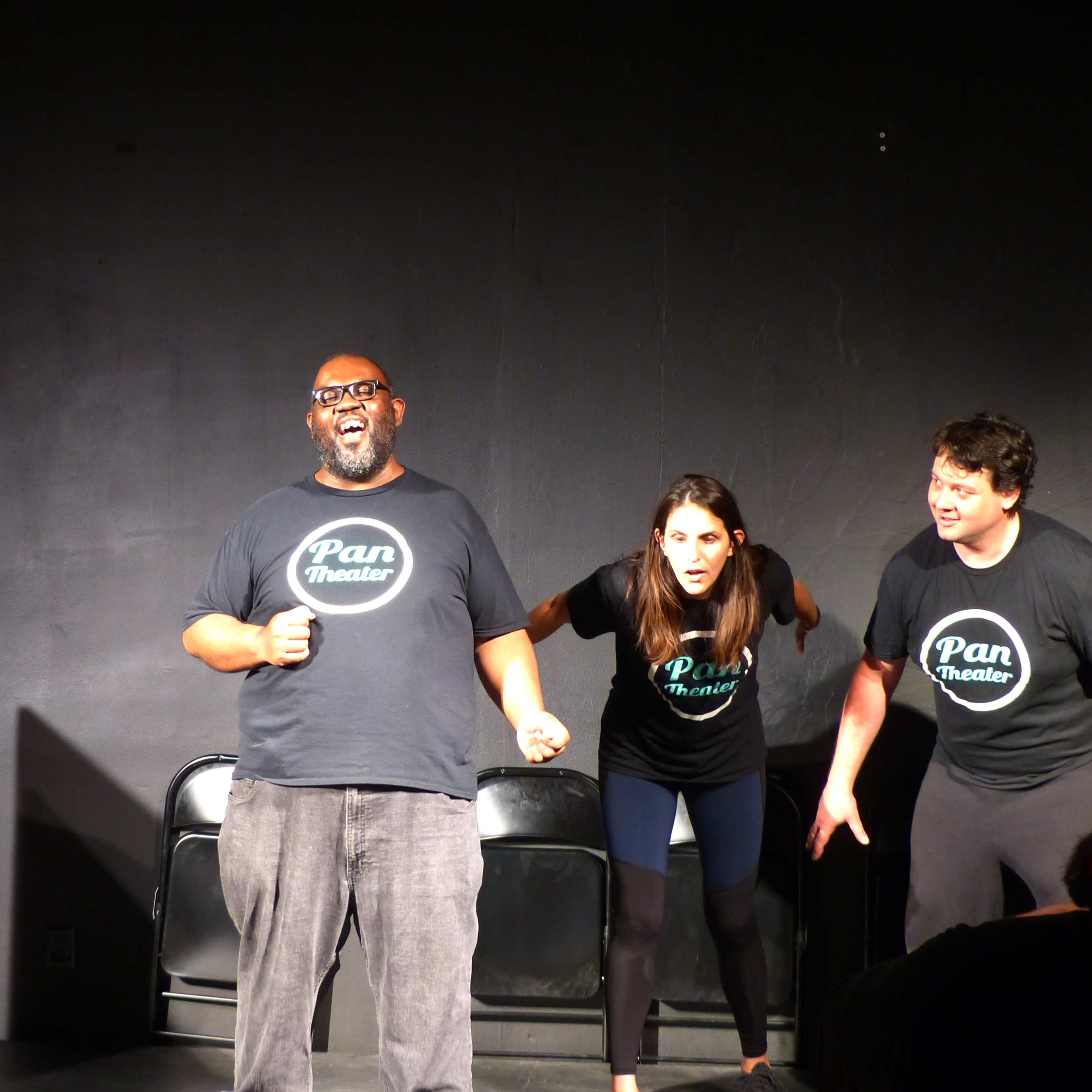 Three performers take the stage in black t-shirts with the "Pan Theater" logo, engaging in an energetic improv scene.