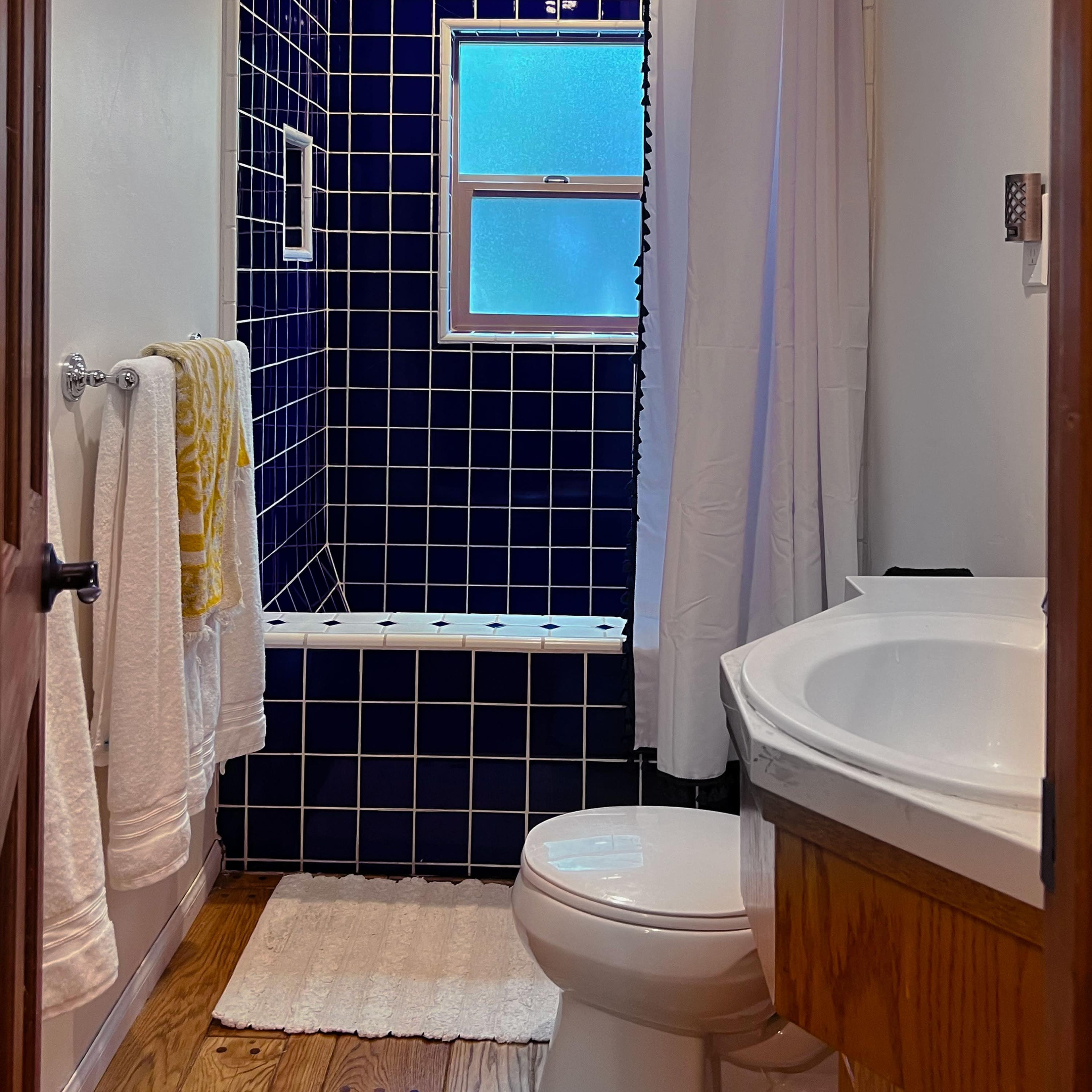 A small bathroom features blue tiled walls, a shower with a curtain, a toilet, and a sink with wooden cabinetry.