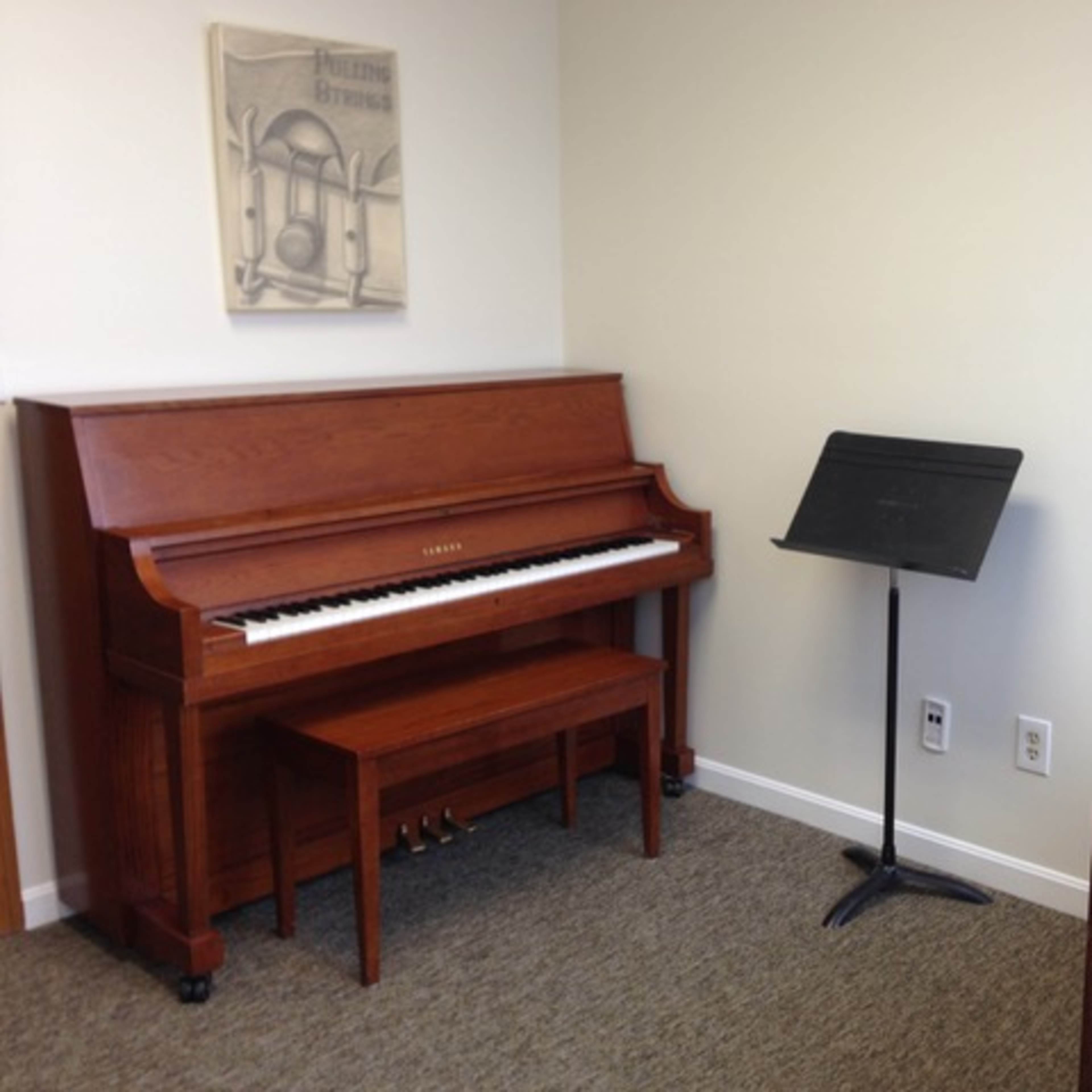 A music room featuring a wooden piano, a music stand, and a small table with a chair against a plain wall.