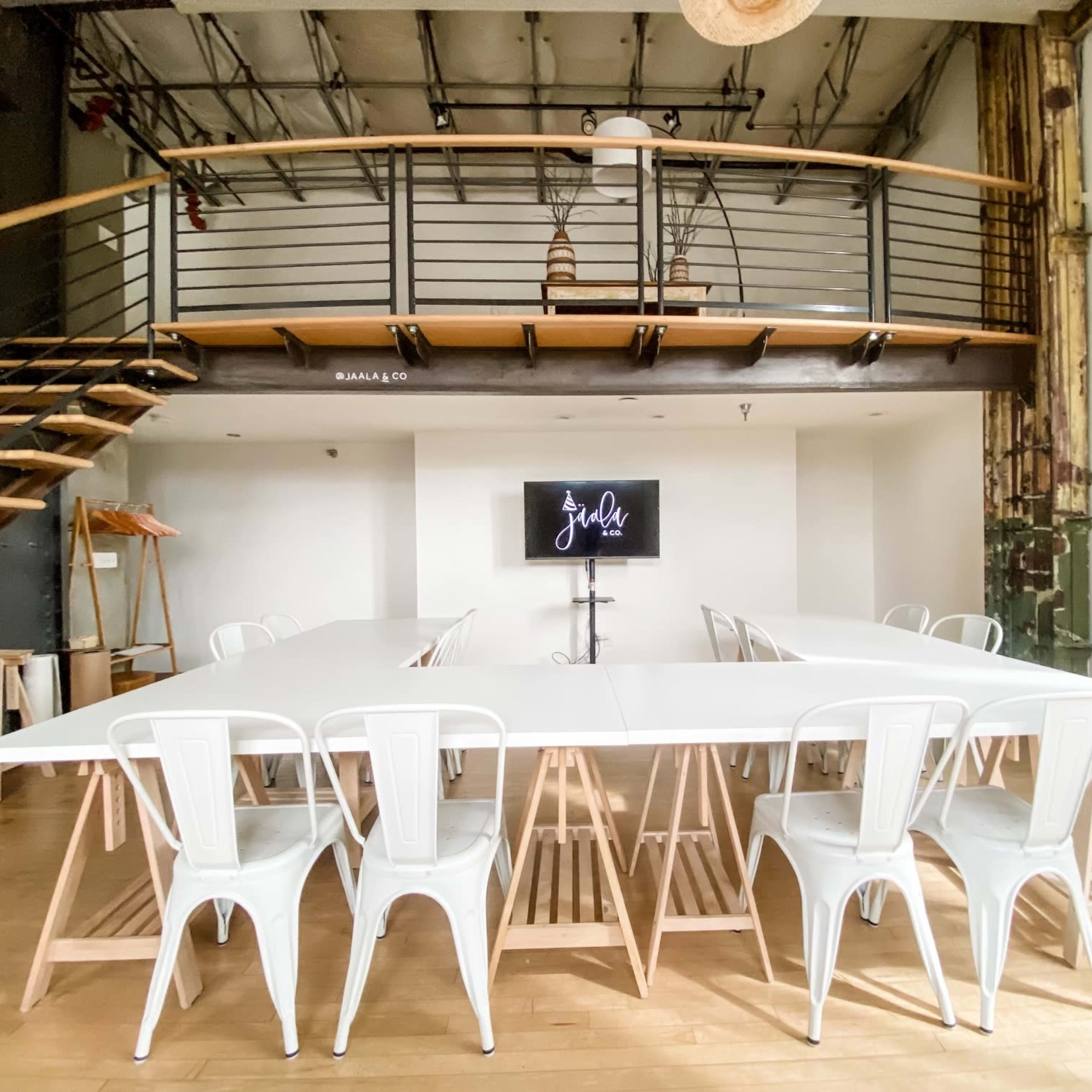 A spacious meeting room with a long white table surrounded by metal chairs, a display screen on a stand, and a staircase leading to a second level.