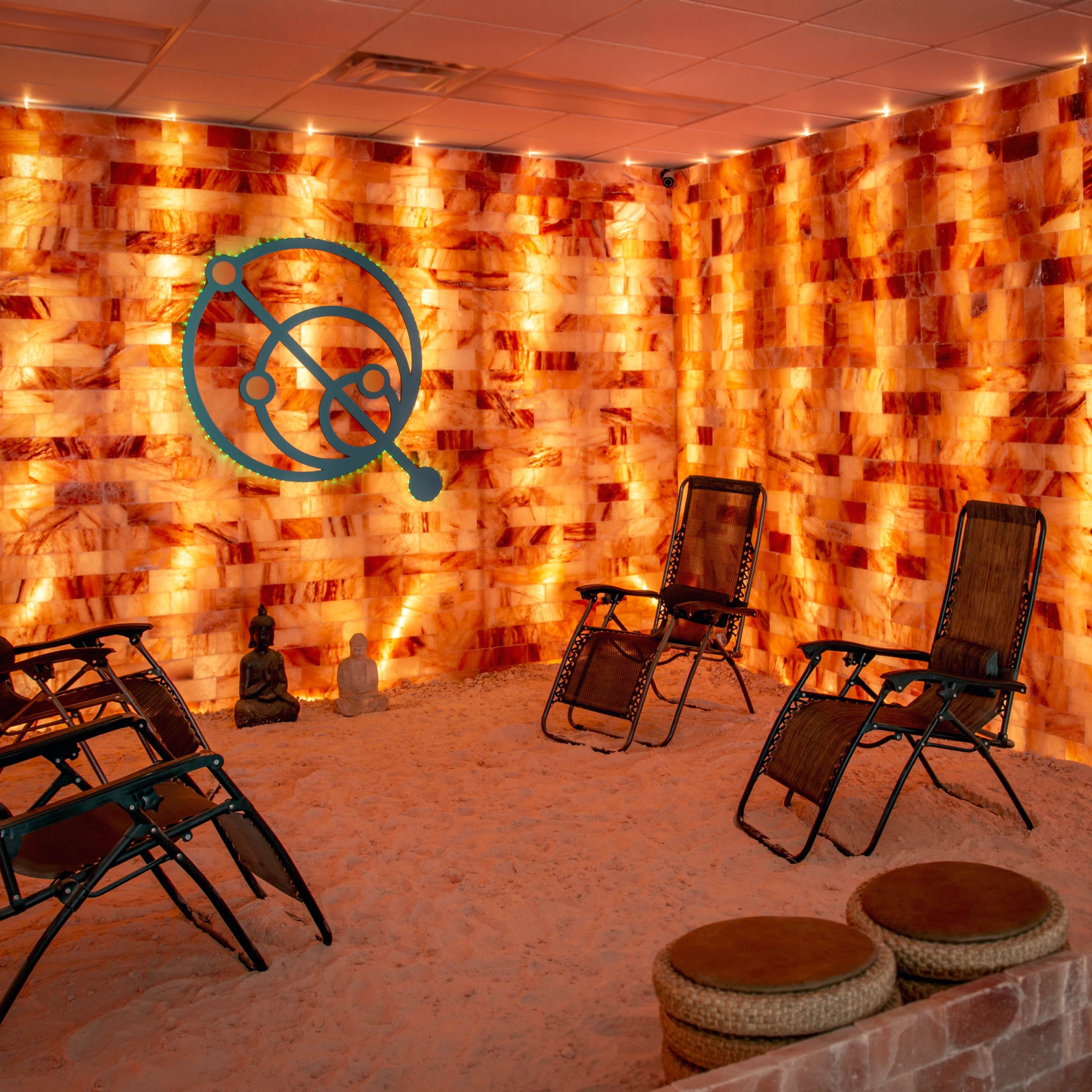 A therapy room with salt brick walls, illuminated by soft lighting, containing several chairs arranged in a circle on sandy flooring.
