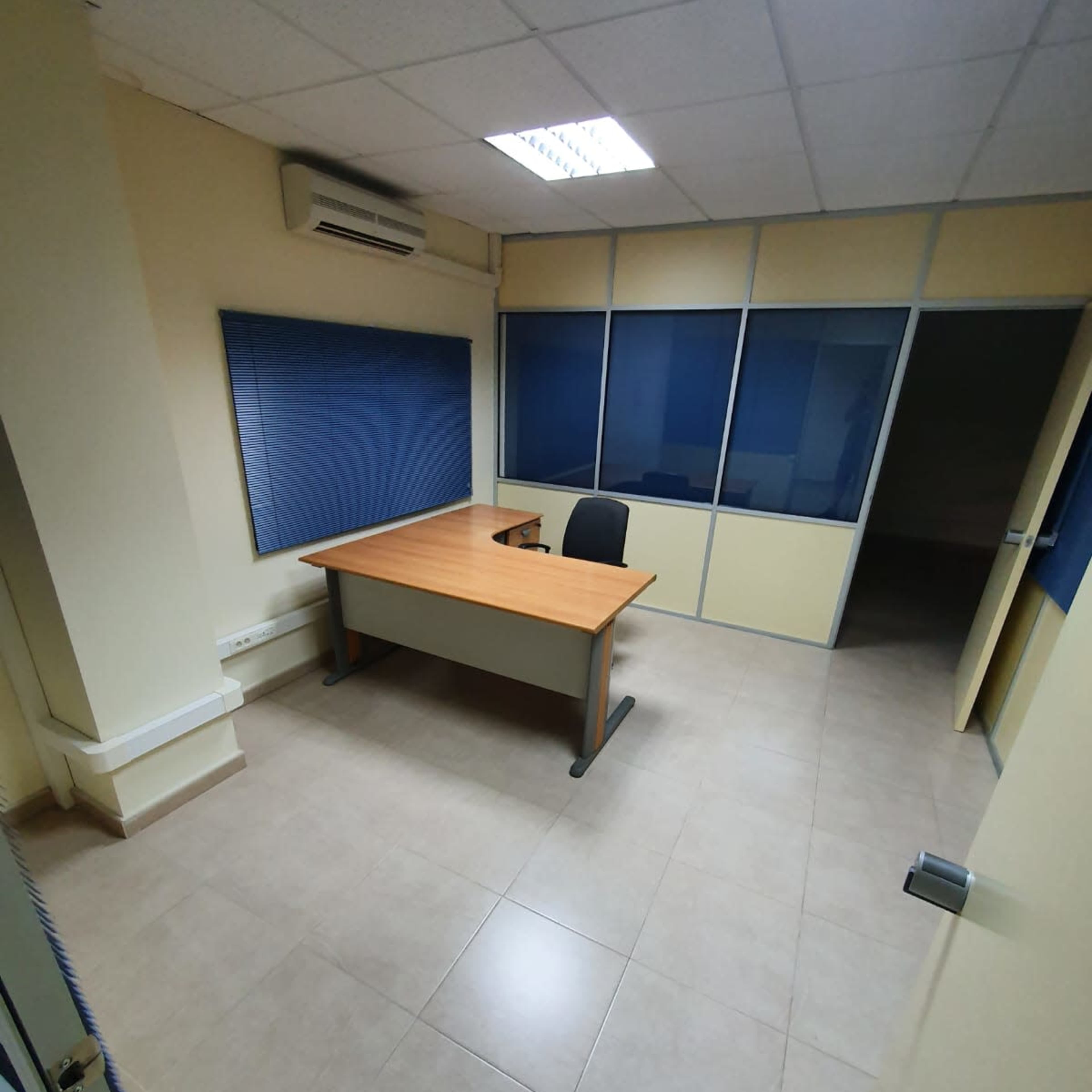An empty office space features a wooden desk and a chair positioned against a window with blue coverings.