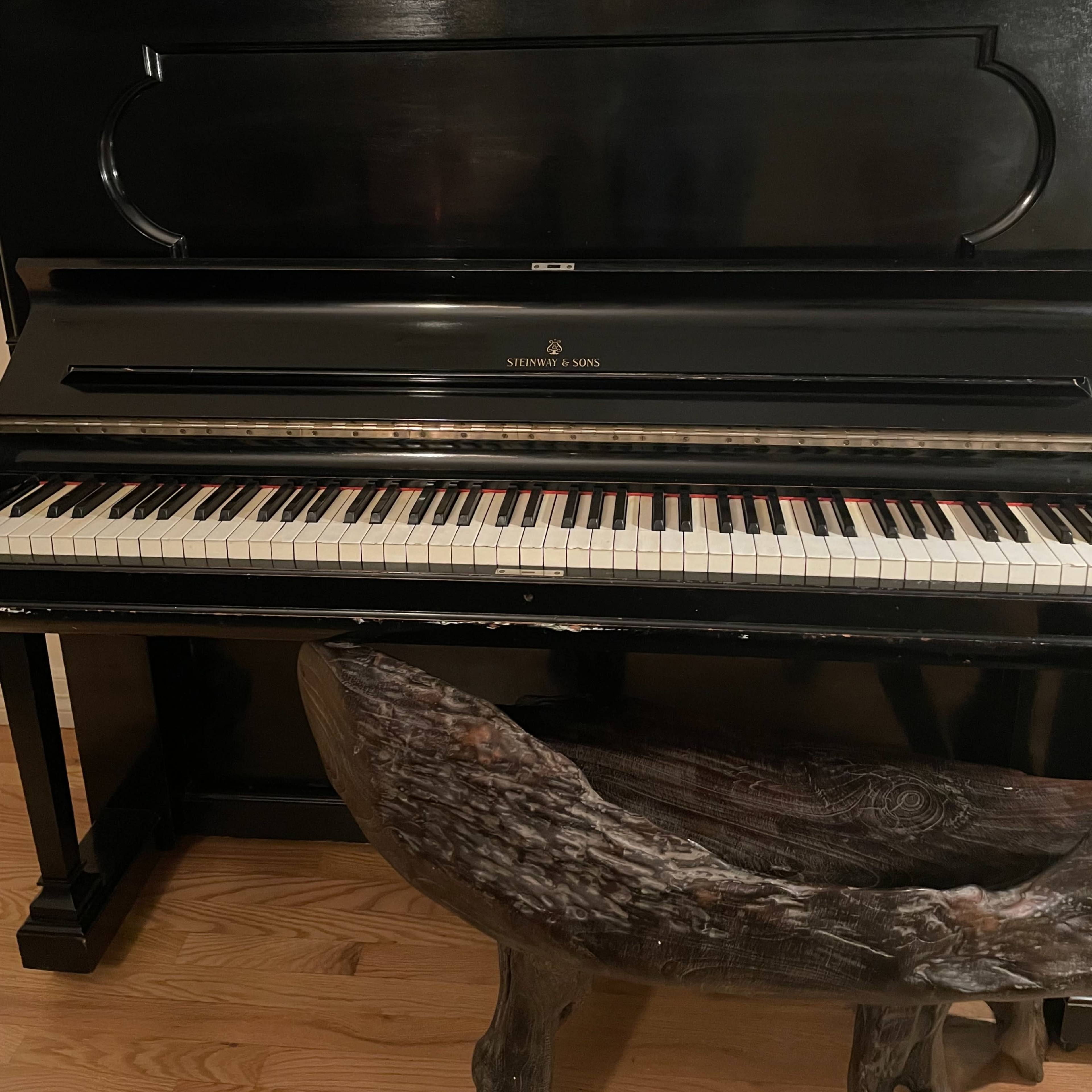A black piano with a wooden bench is positioned beneath a framed artwork, accompanied by a white vase and a candle holder on a wooden floor.