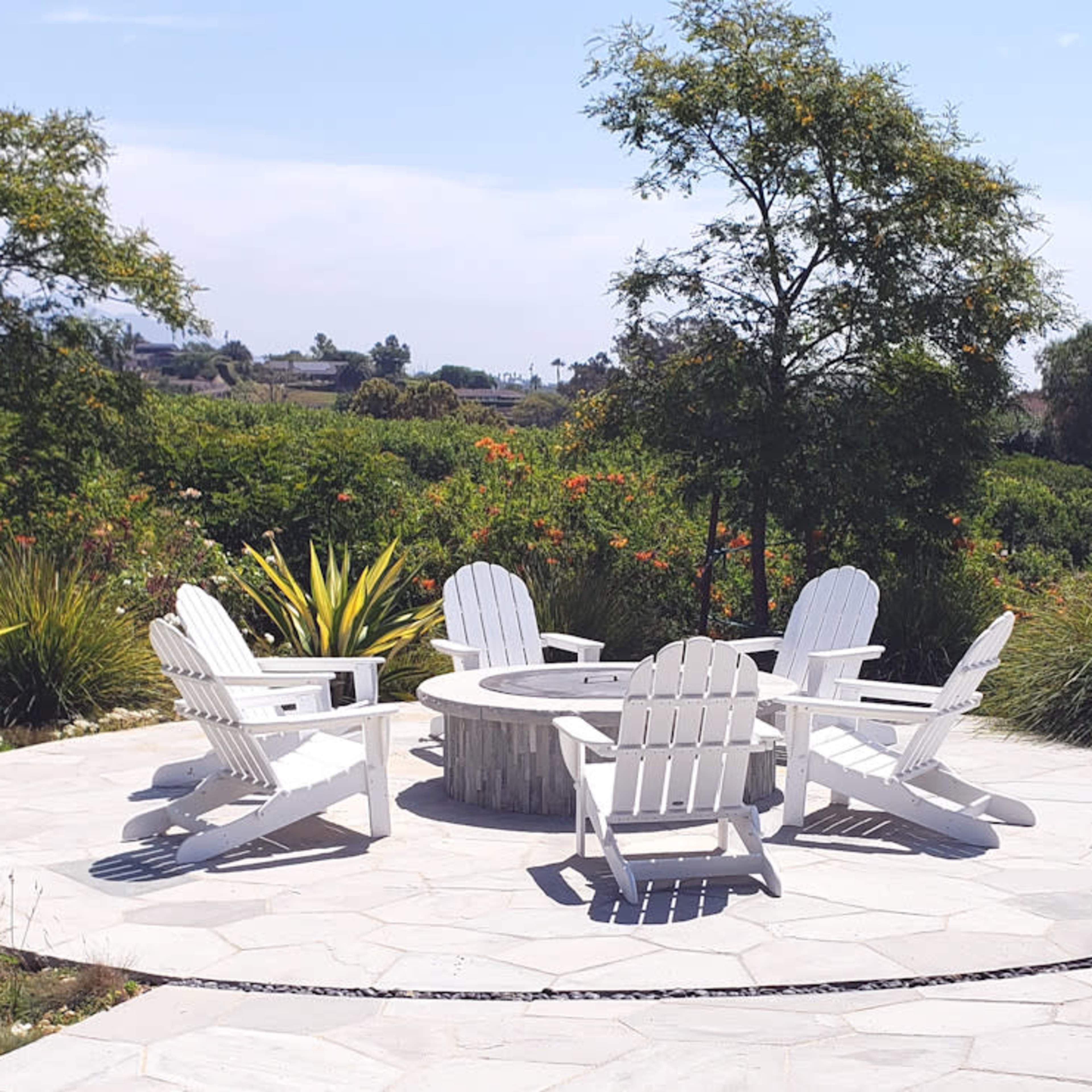 A circular stone patio features five white Adirondack chairs arranged around a fire pit, surrounded by greenery and trees.