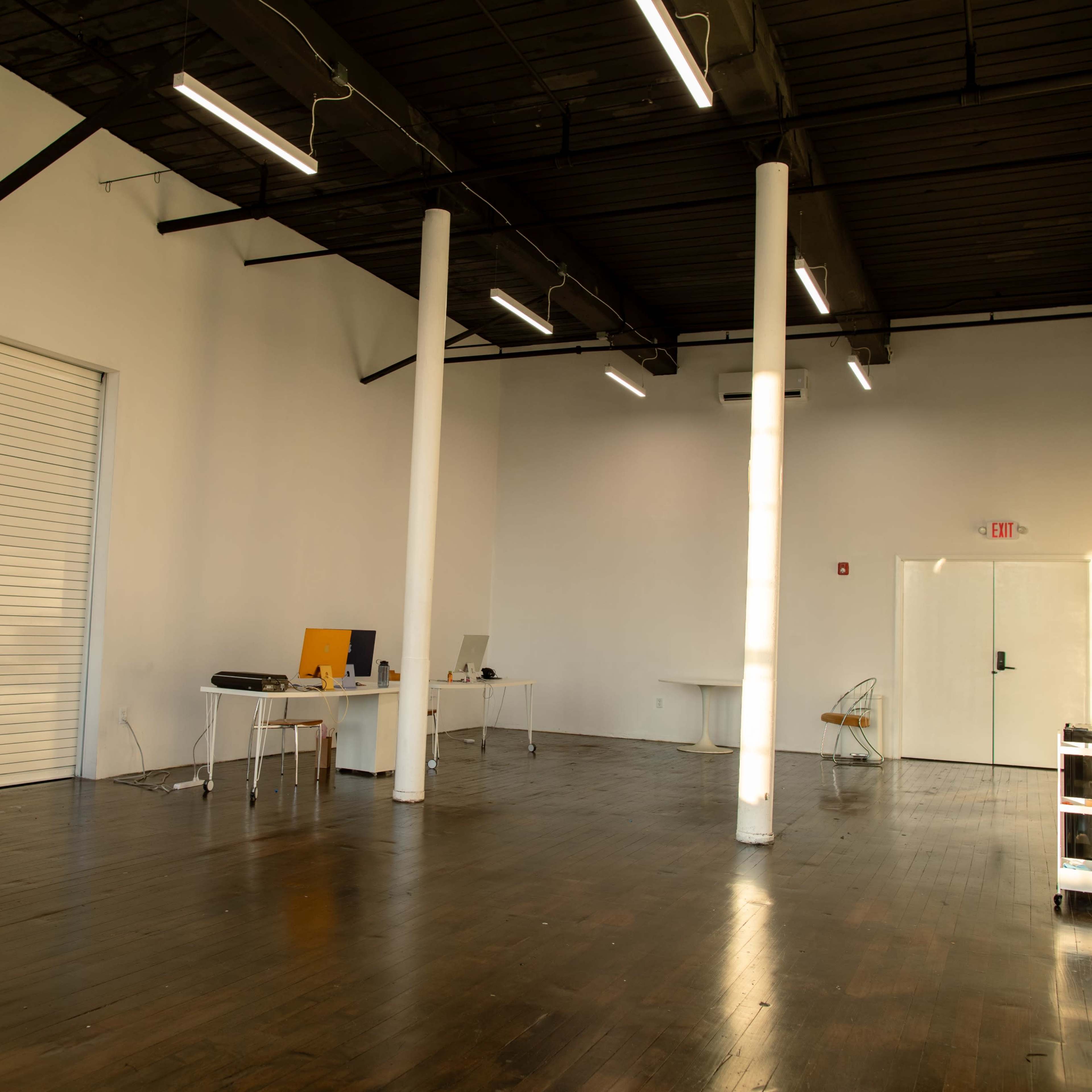 A spacious, empty interior of a modern office or studio with wooden floors and white walls, featuring several tables and chairs, along with a closed overhead door.