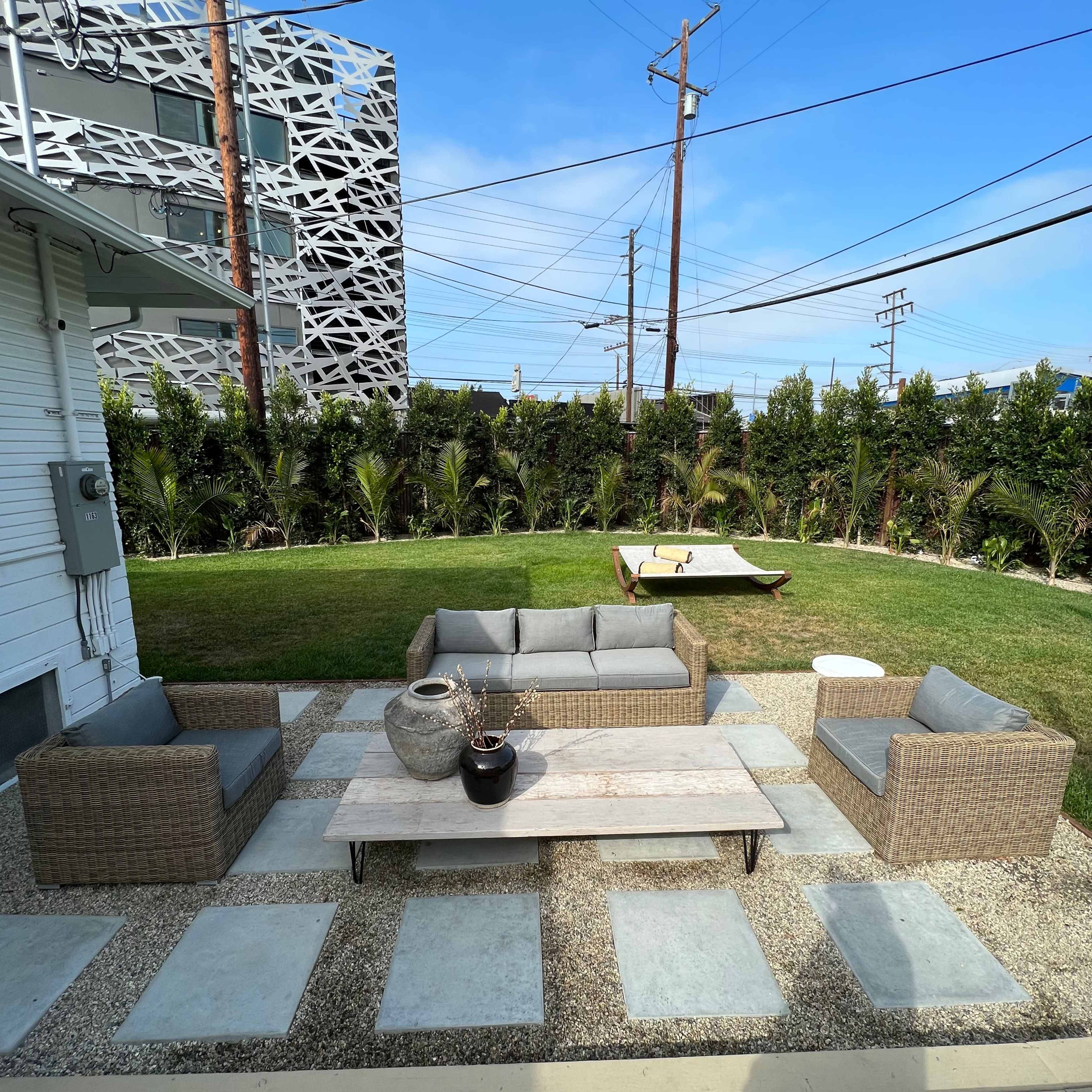 An outdoor seating area with wicker furniture arranged around a stone table, set on a gravel surface with concrete stepping stones, and a lawn in the background.