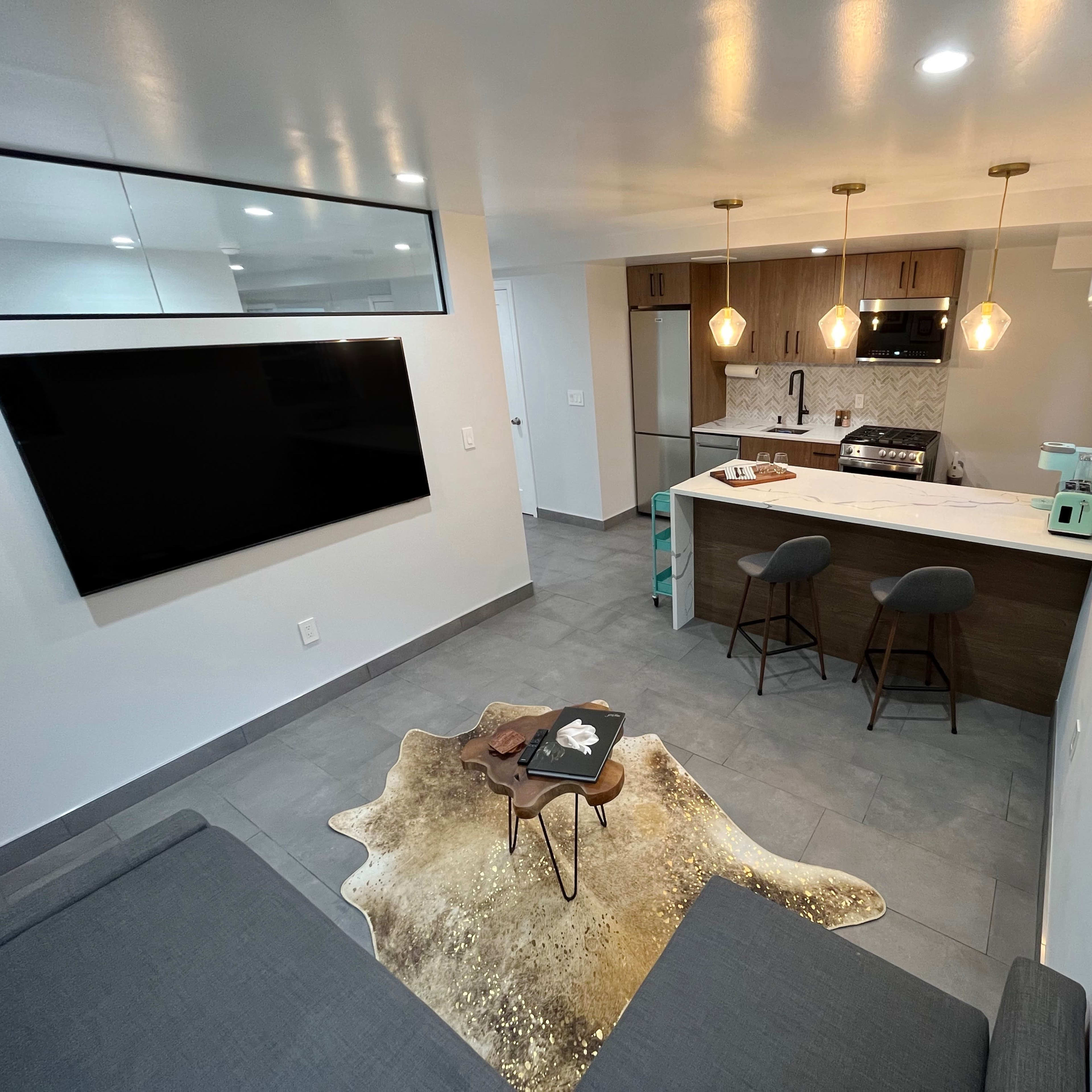 A modern living space with a kitchen area featuring bar stools, a television mounted on the wall, and a decorative rug on the floor.