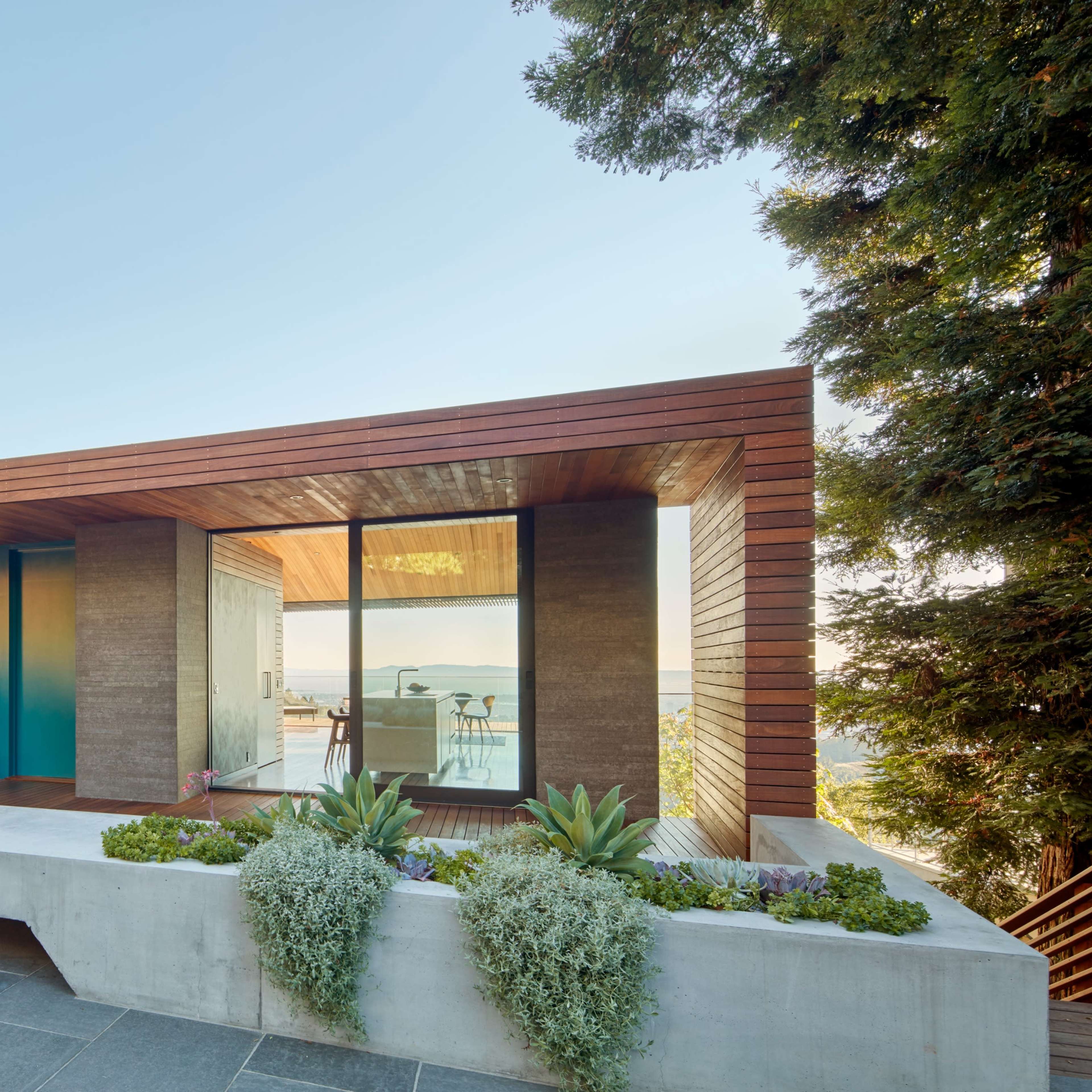 A modern, single-story house with a wooden overhang, large glass windows, and a landscaped front garden.